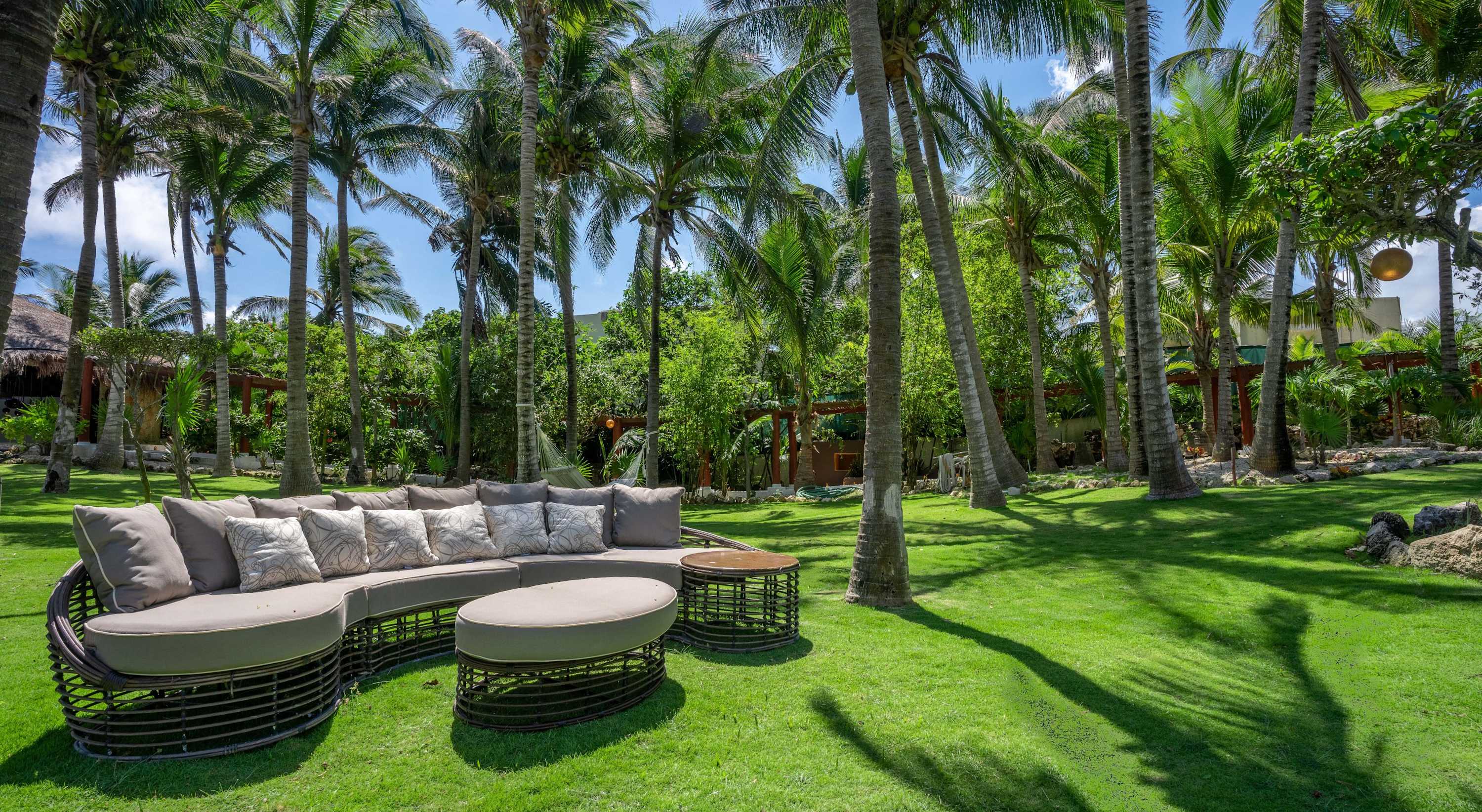 A curved outdoor sofa and ottoman with patterned cushions sit on a lush green lawn surrounded by pal