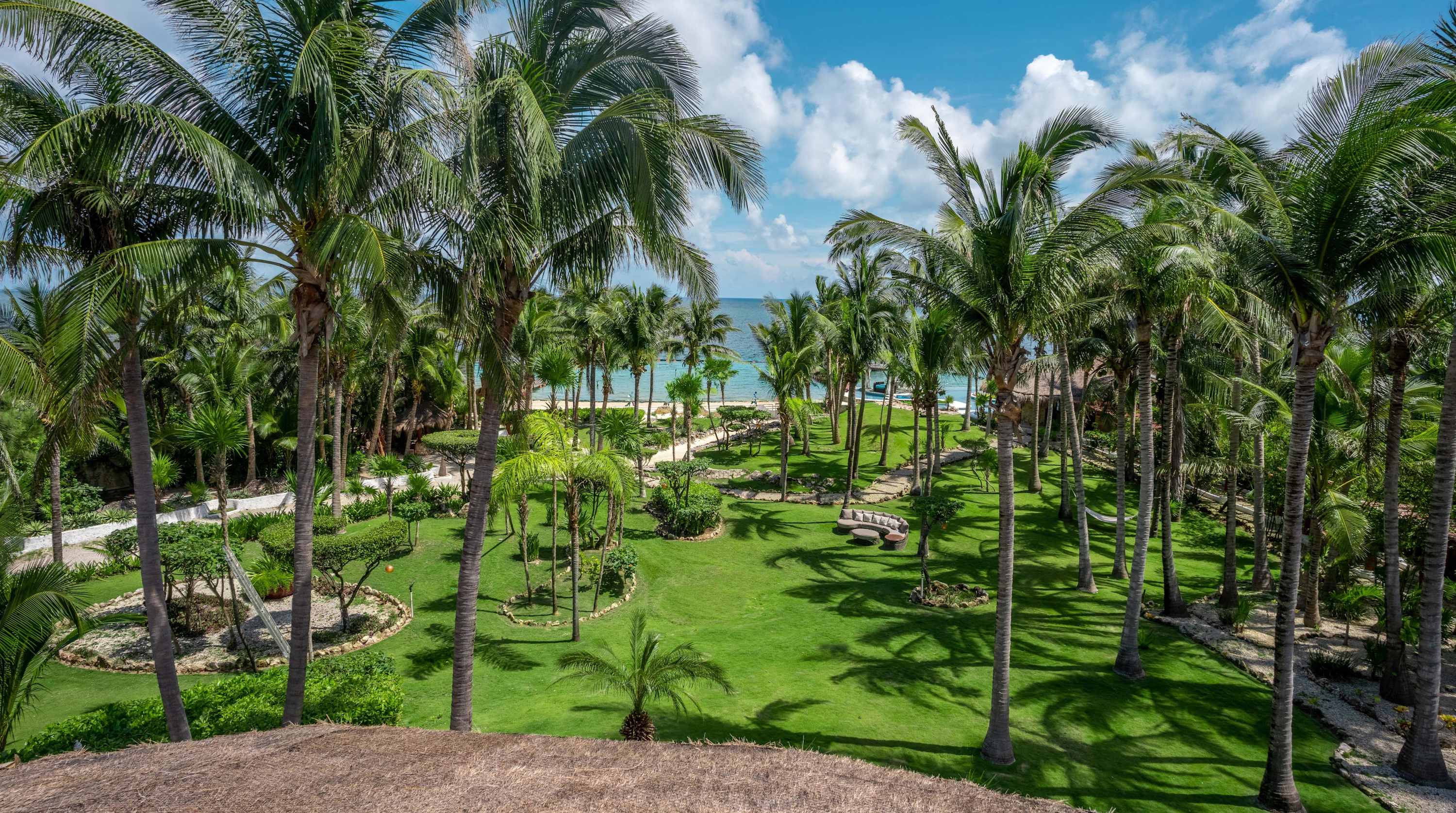 Lush palm trees and green lawns lead to a tropical beach under a blue, cloudy sky.