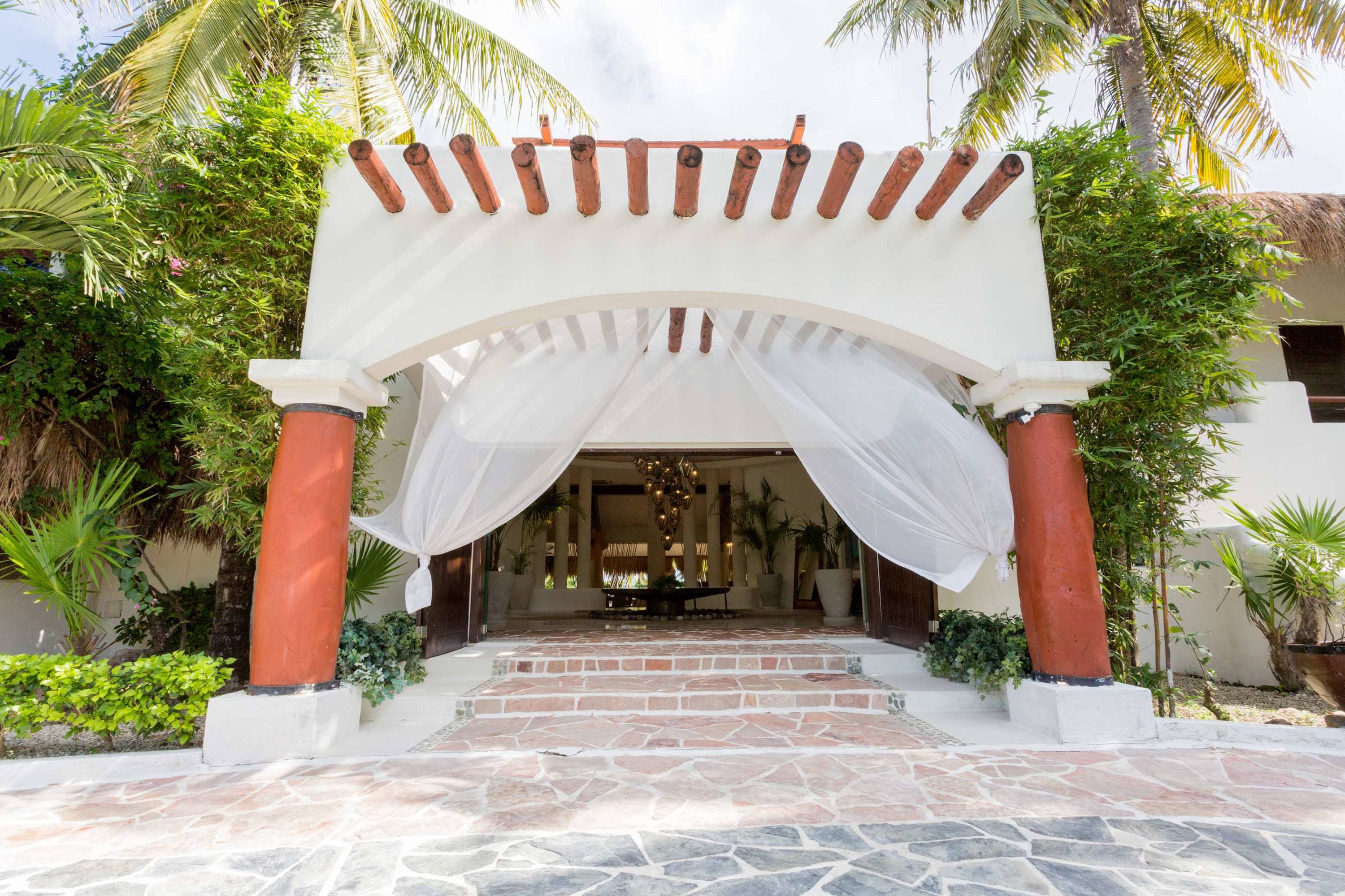 Entrance to a building with an archway and white drapery, surrounded by tropical plants and palm tre