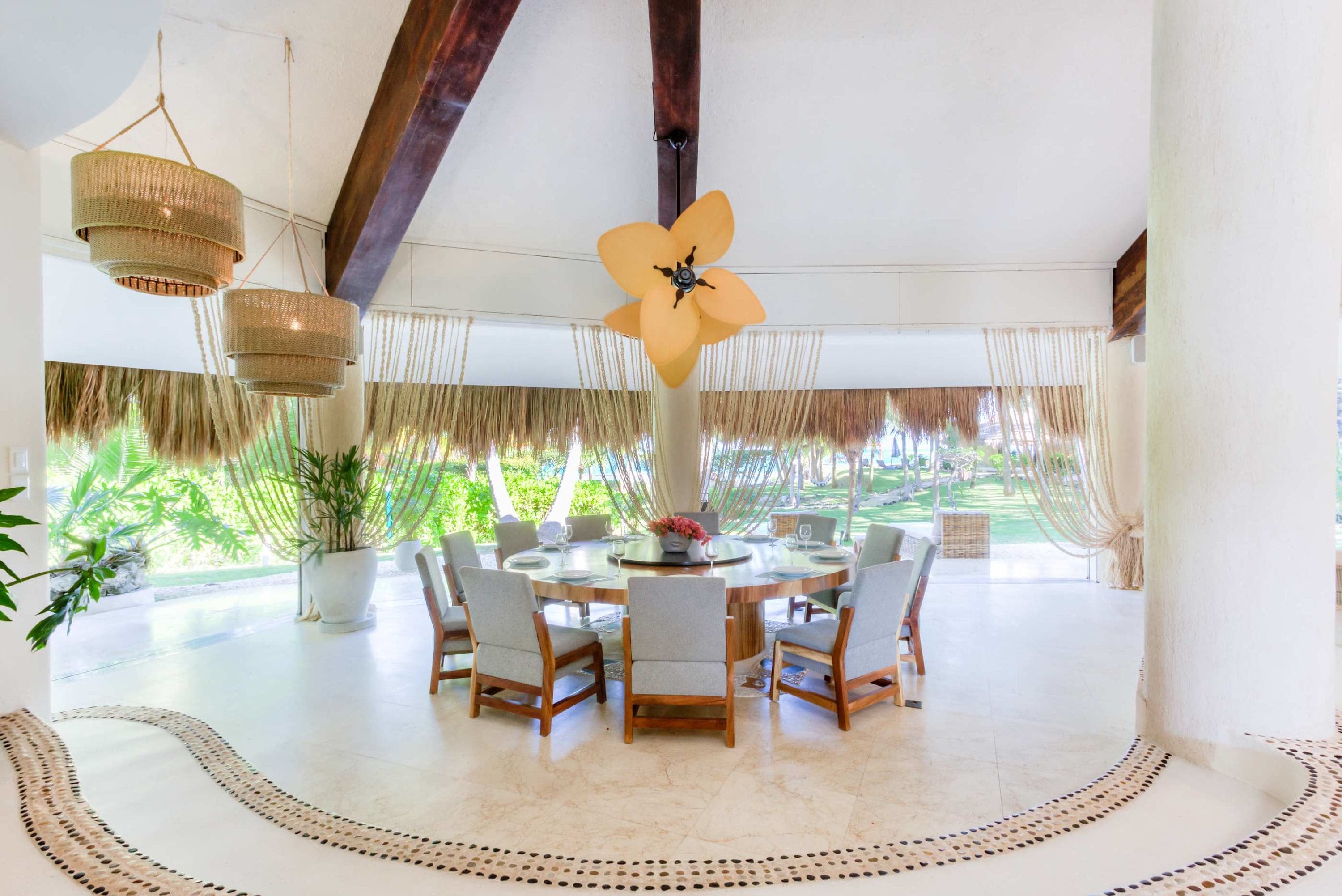 Tropical dining room with thatched roof curtains, rattan light fixtures, and a large wooden table.