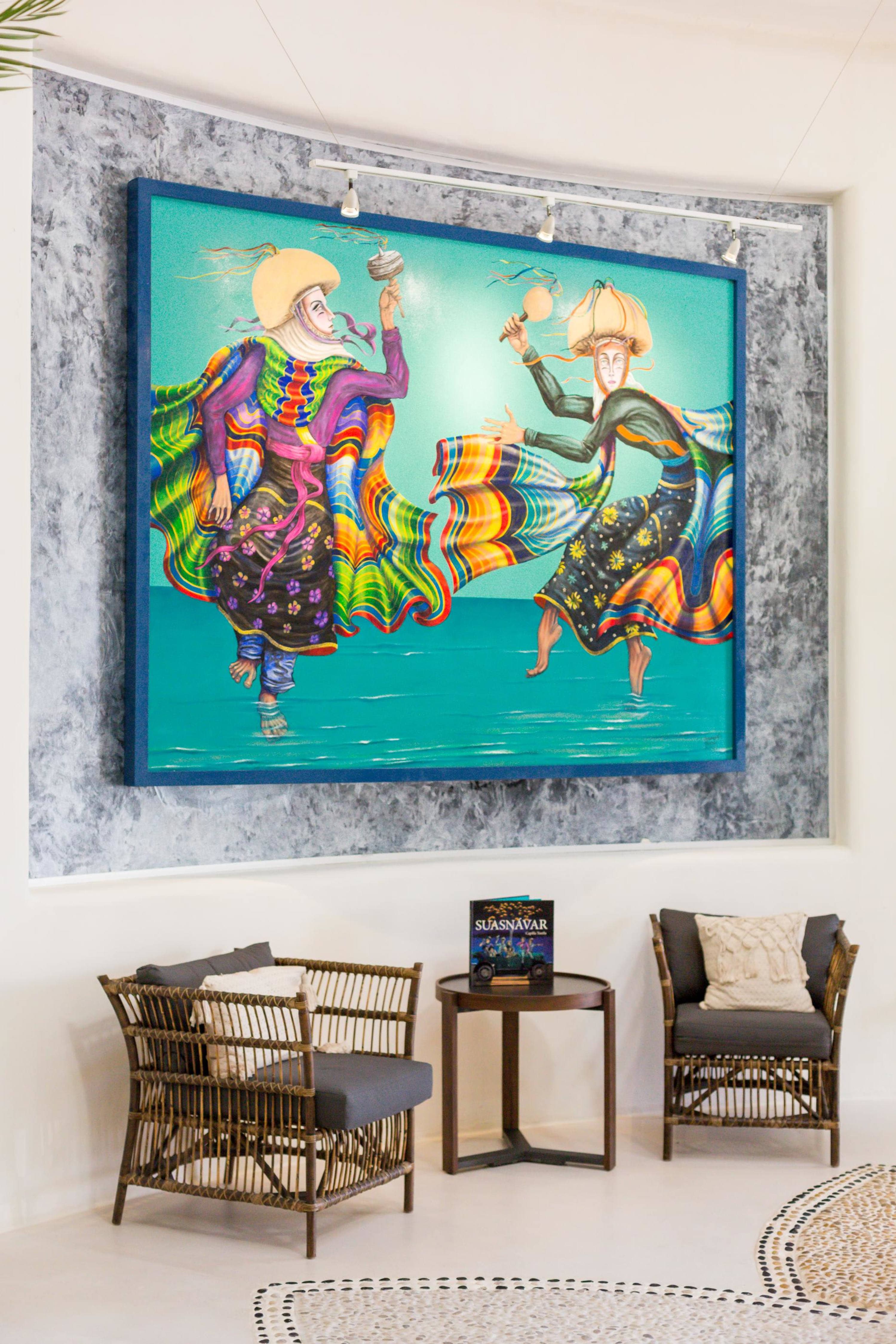 Two figures in colorful costumes dance next to a large painting and two wicker chairs.
