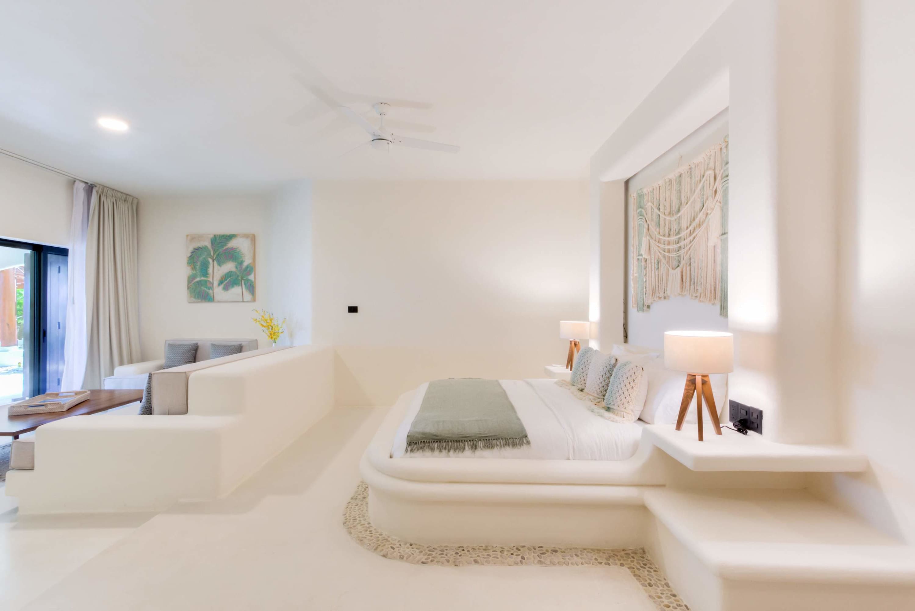 A bright white bedroom with a minimalist design. Features a low-profile bed with a green throw blank