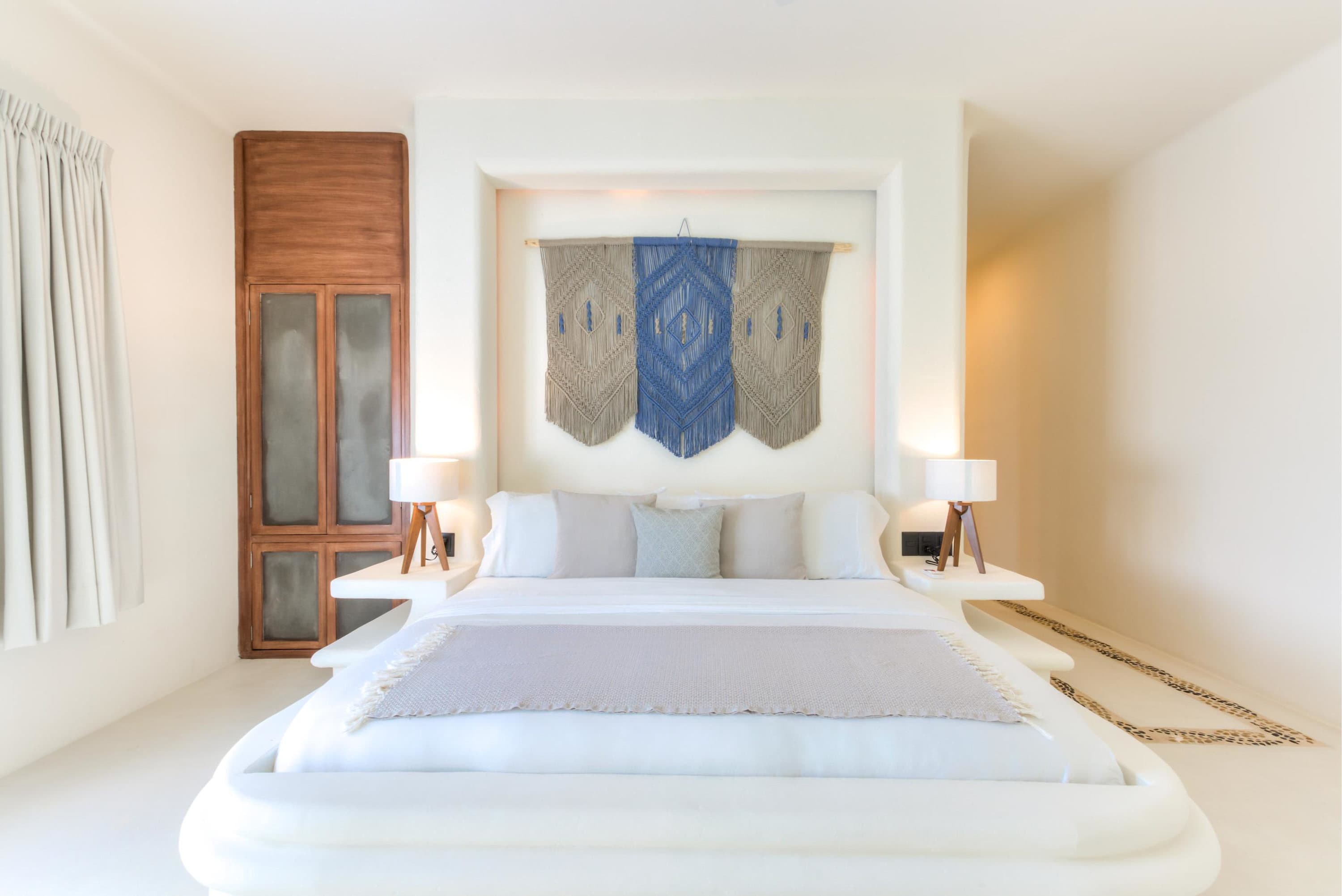 A modern bedroom with a white bed, macrame wall hanging, and wooden closet.
