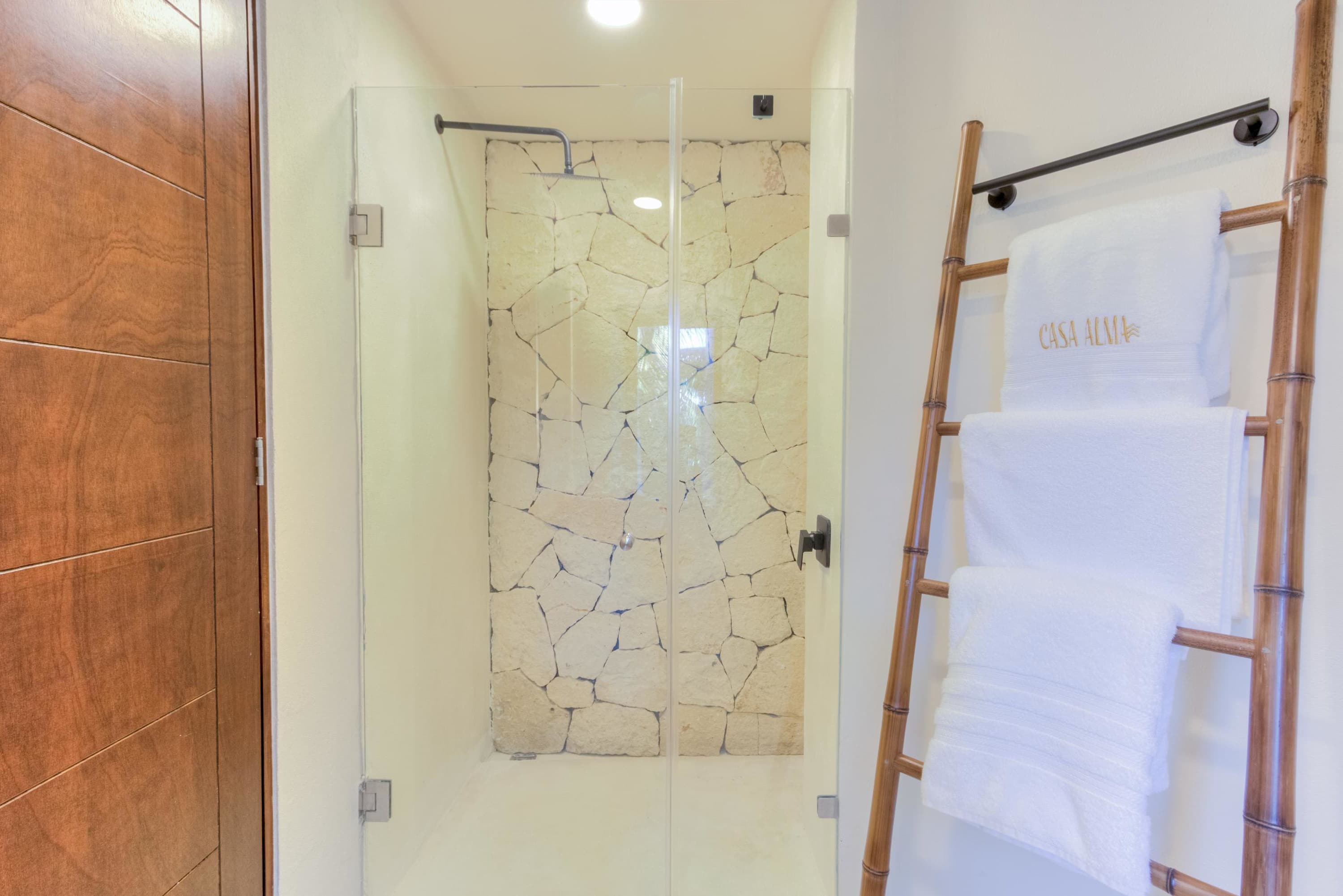 A modern bathroom with a glass-enclosed shower, stone walls, and a bamboo towel rack.