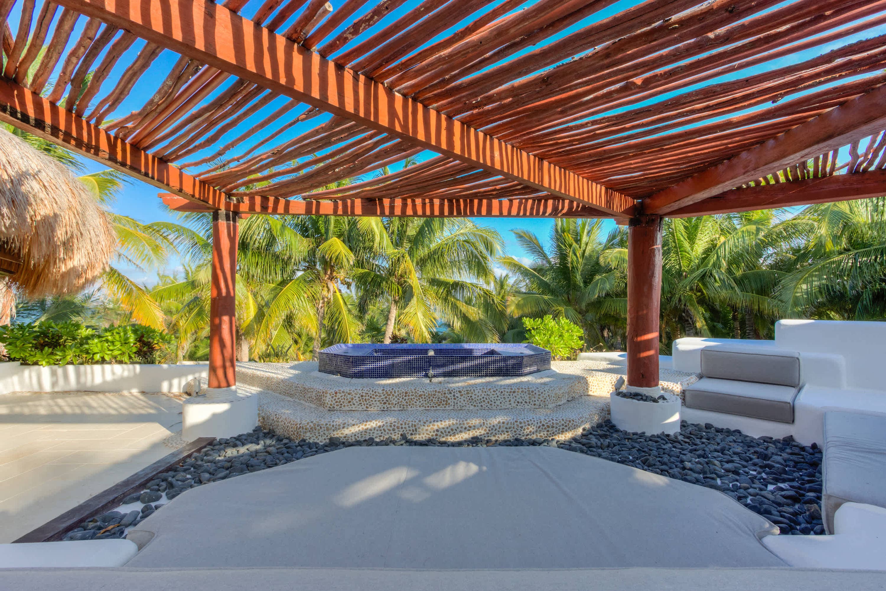 A thatched roof pergola shades a hot tub surrounded by palm trees and a seating area.