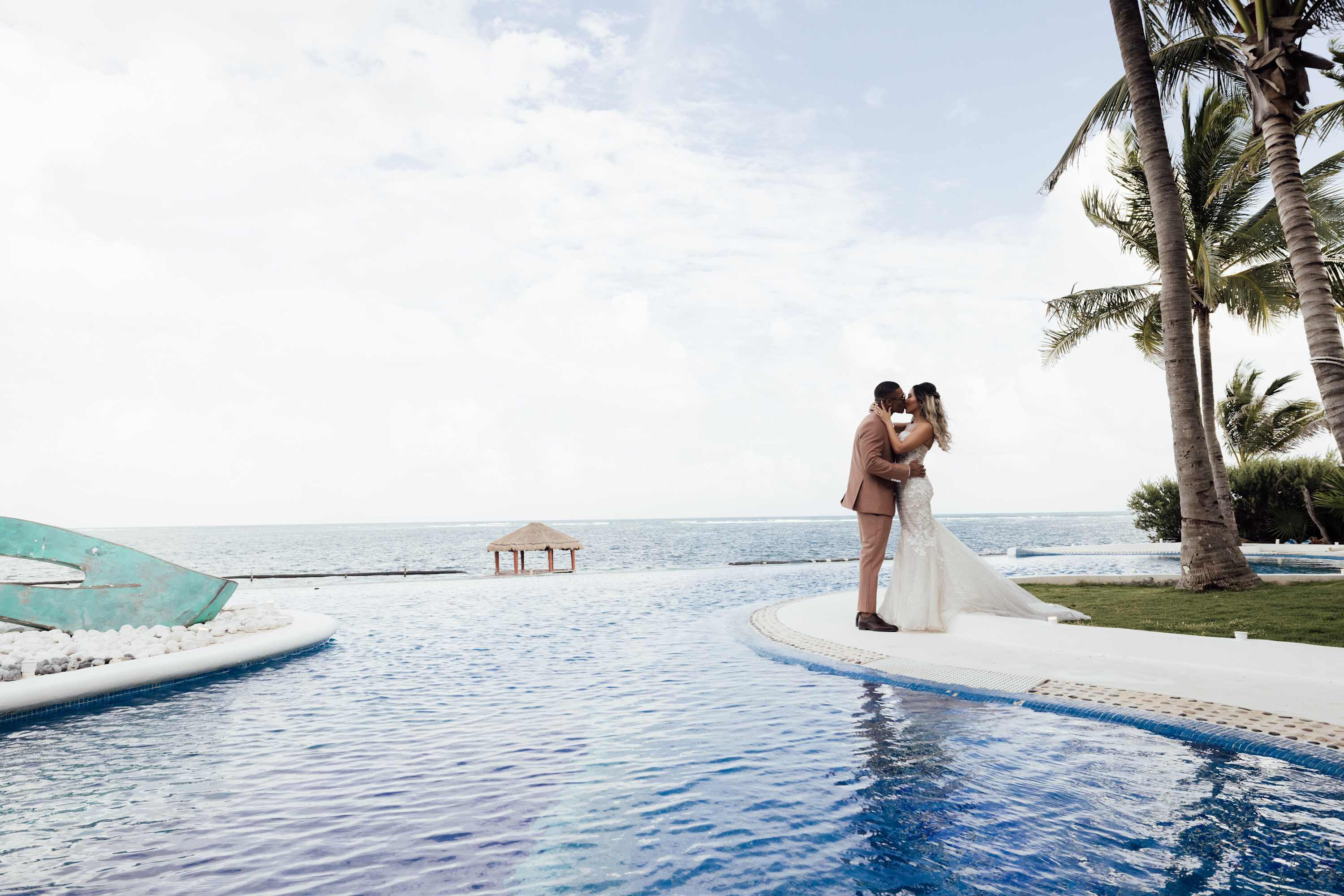 A bride and groom kiss by a pool with the ocean and palm trees in the background.