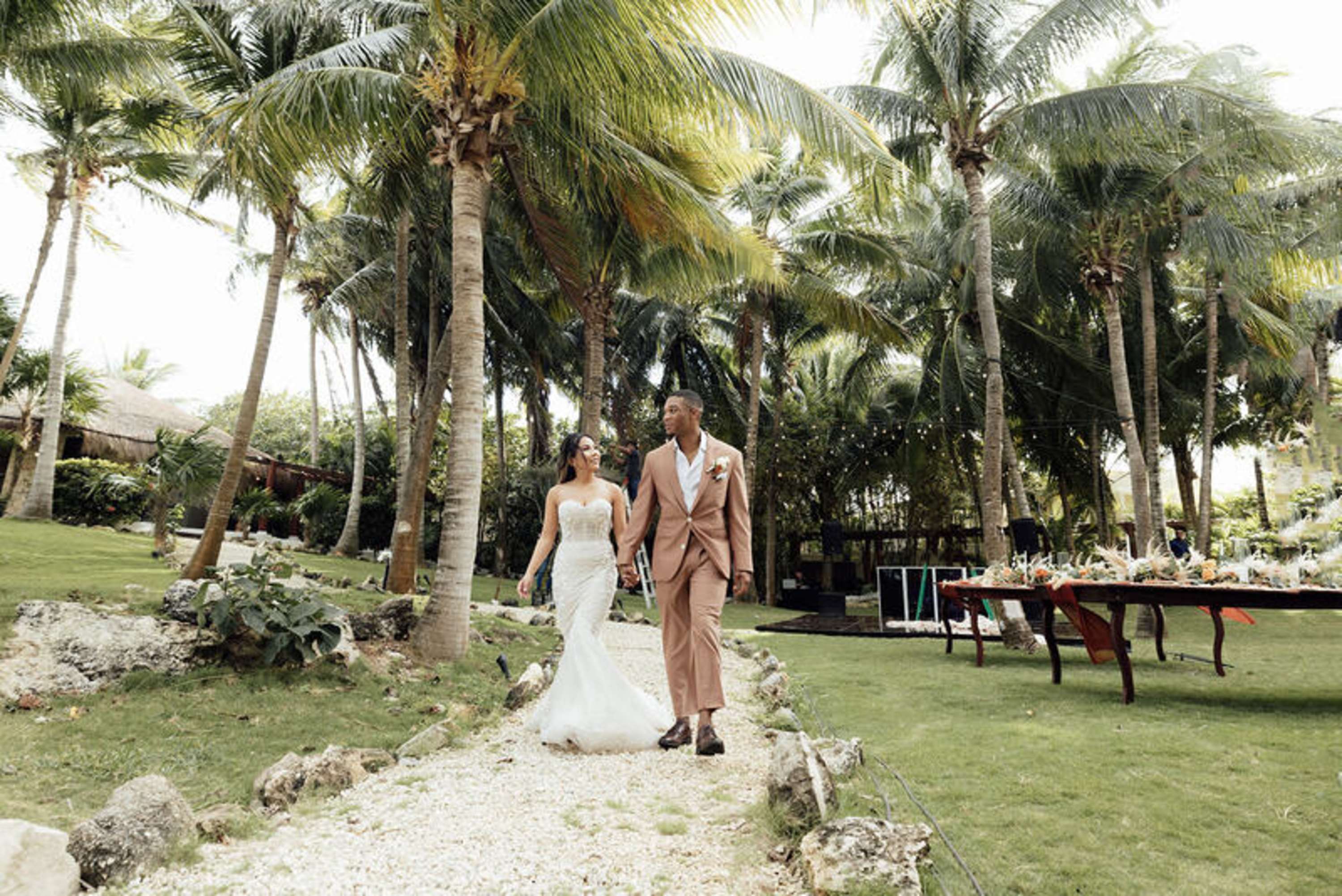 A bride and groom walk hand-in-hand down a path surrounded by palm trees.