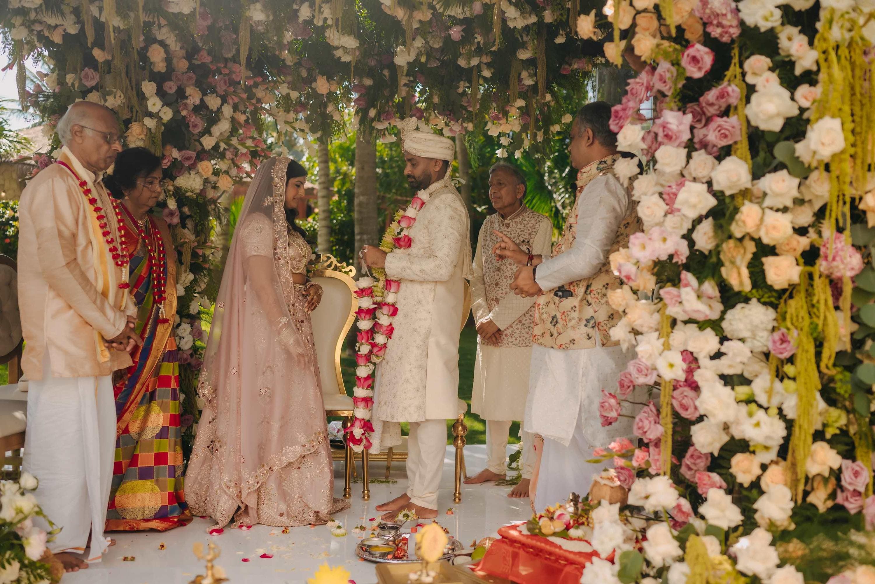 Indian wedding ceremony with couple exchanging garlands under flower arch.