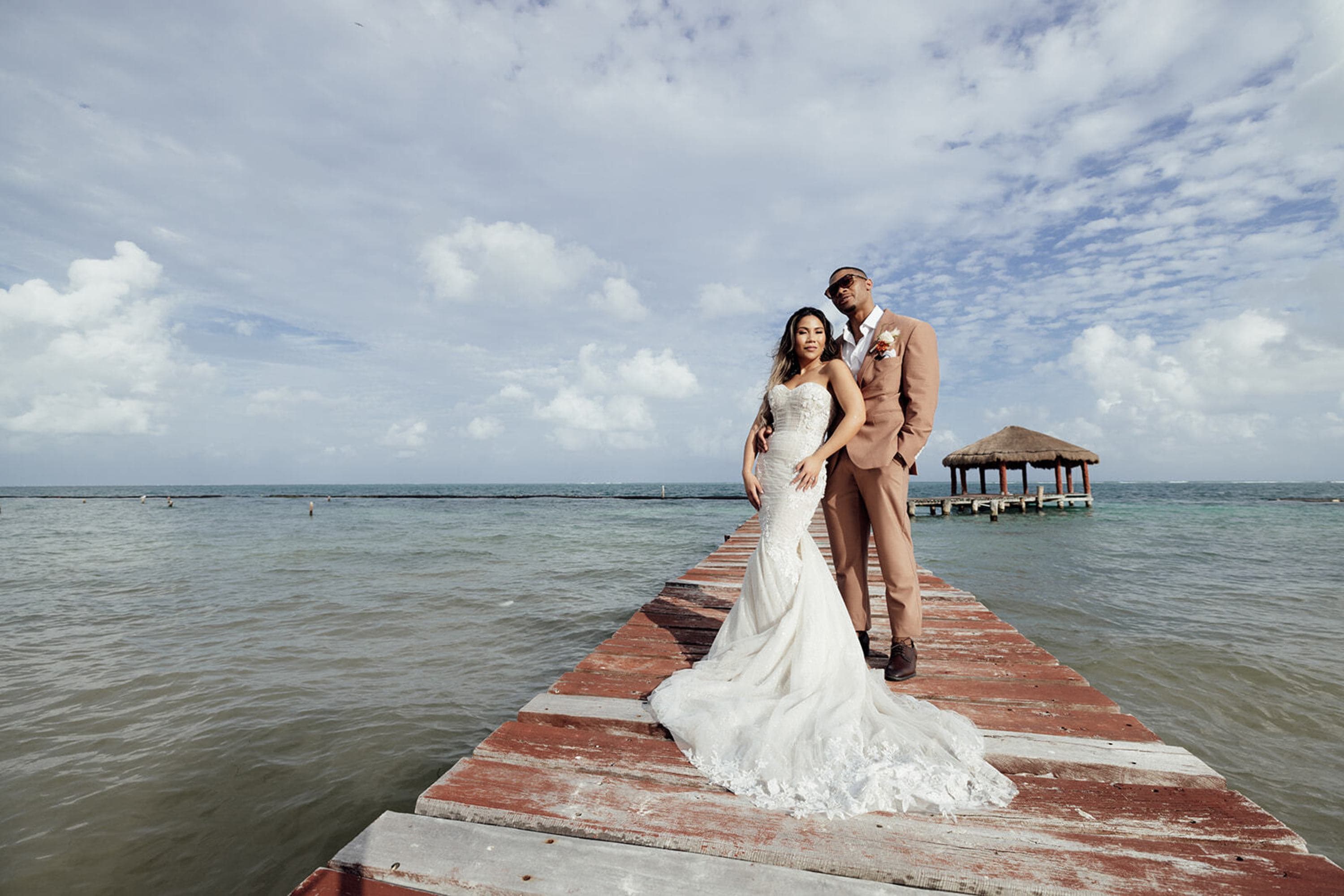 A newlywed couple poses on a wooden pier in front of a thatched-roof gazebo.