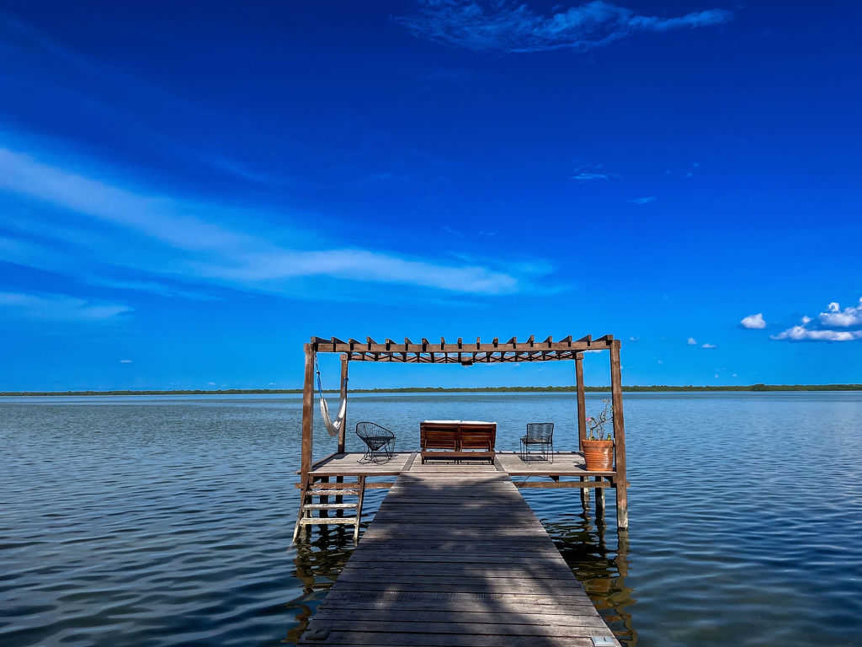 Wooden pier with furniture and a hammock extends into calm water under a bright blue sky.