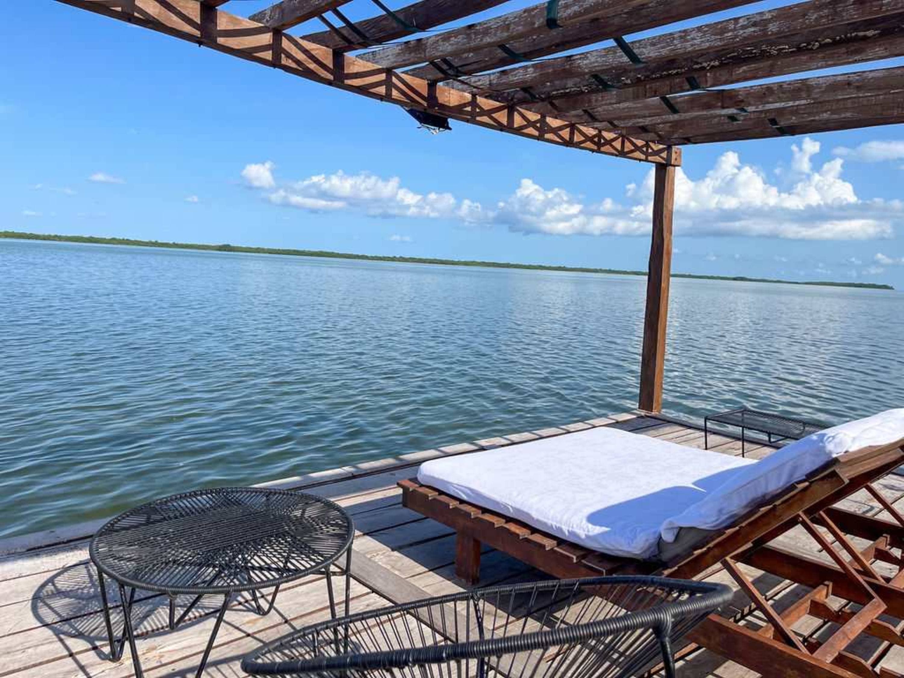 A wooden deck with lounge chairs and a table sits on the water, under a canopy.