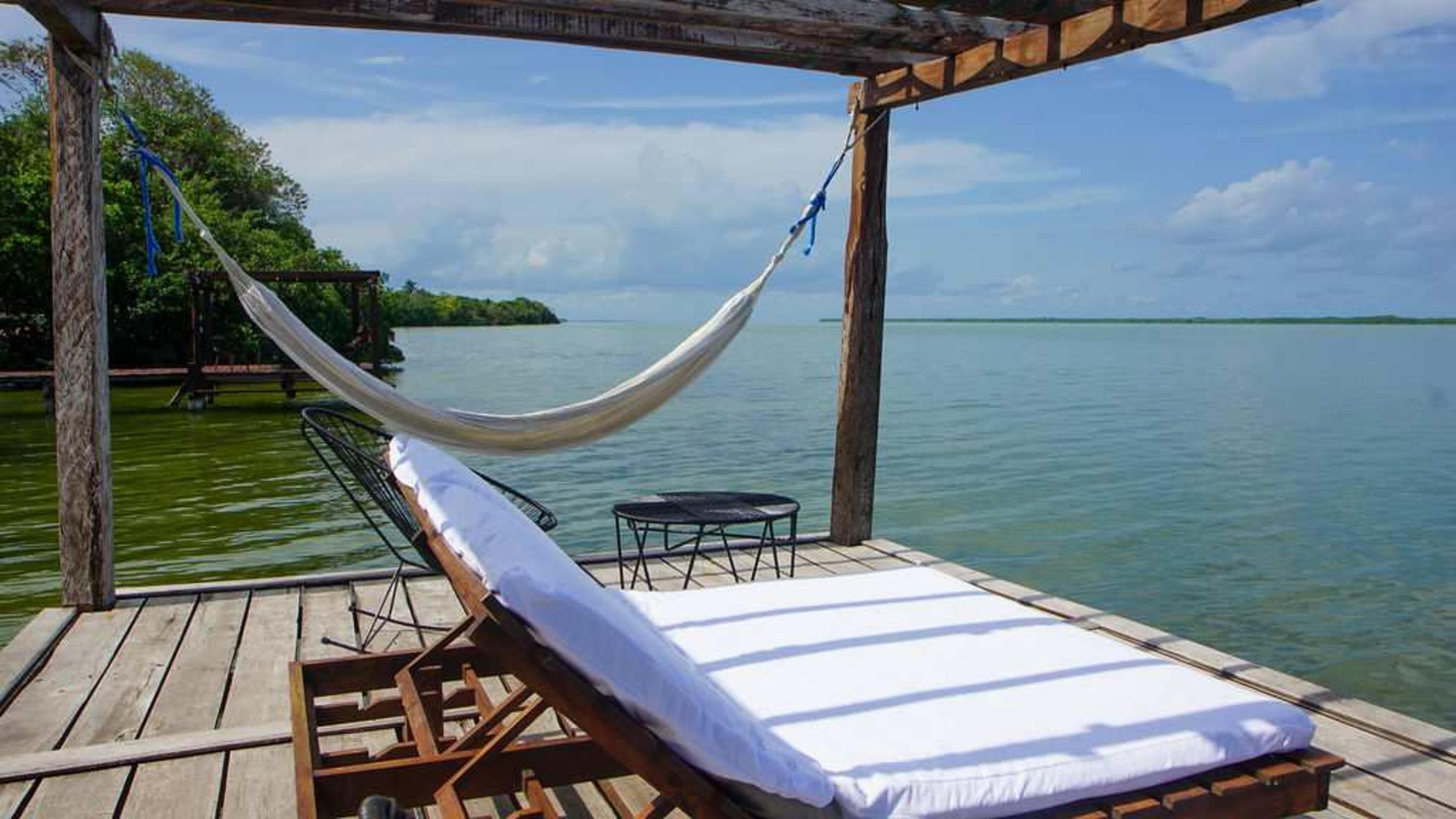 A hammock and lounge chair on a wooden deck overlooking a calm body of water.