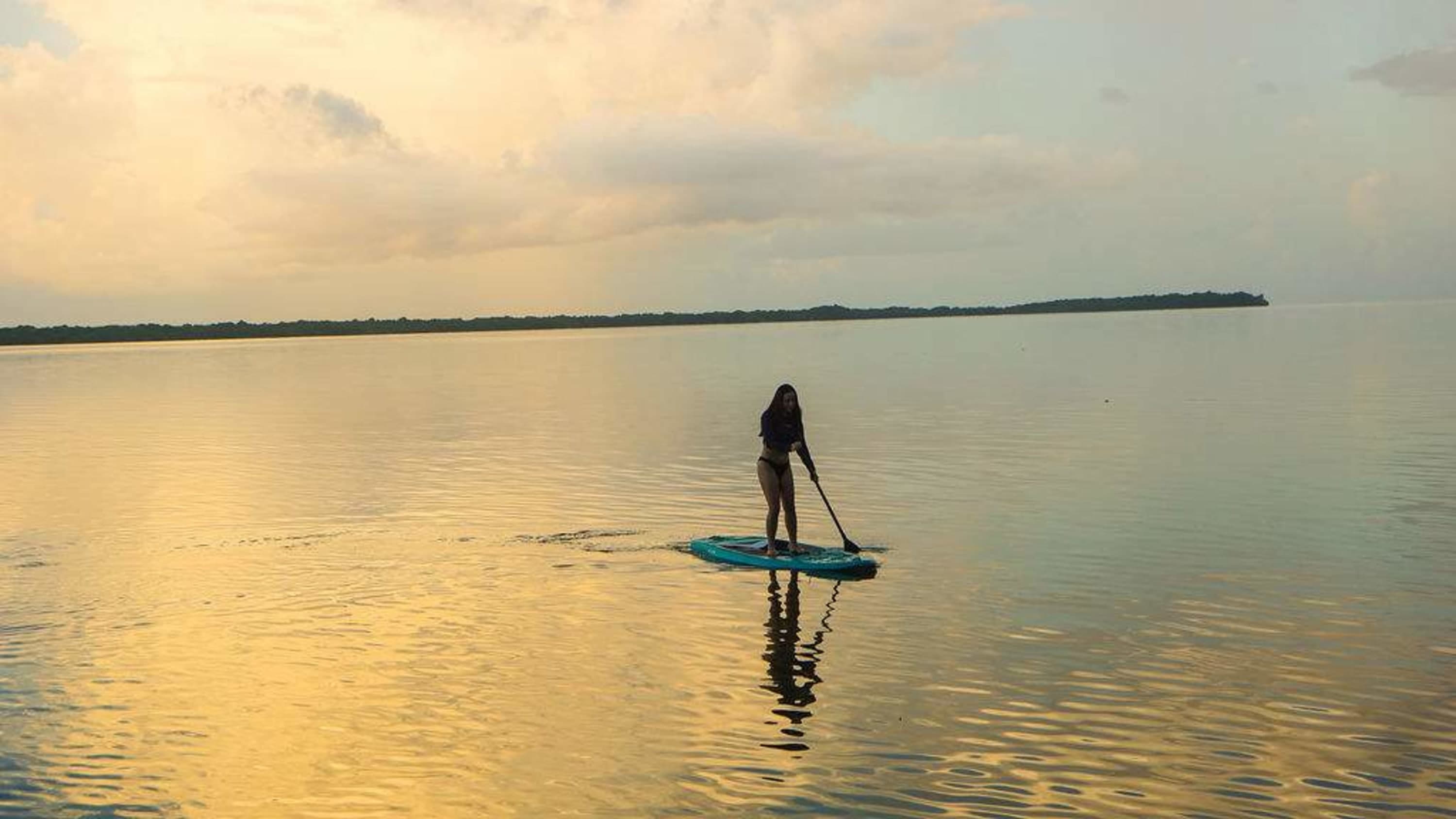 A woman paddleboards on a calm body of water at sunset.