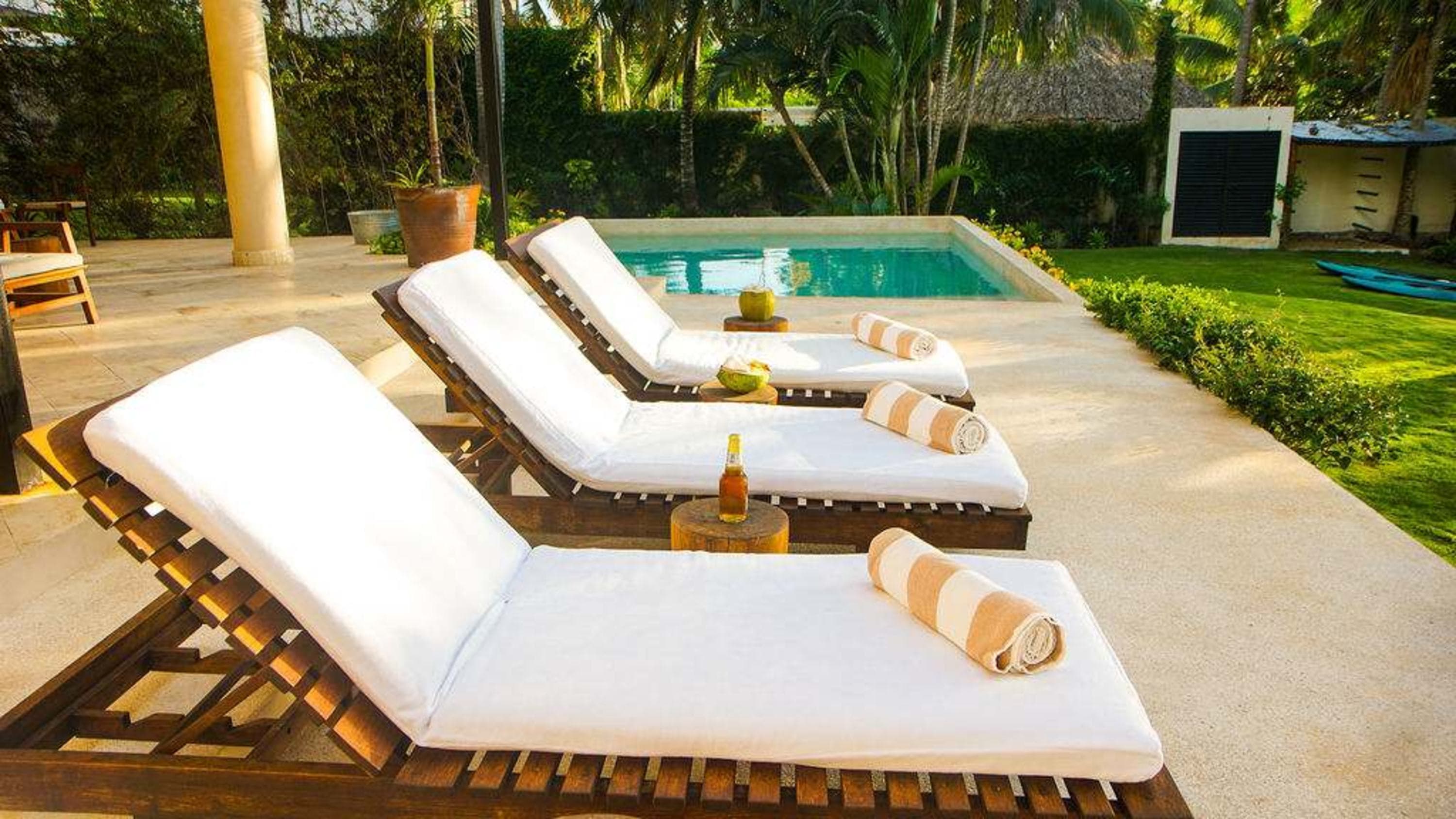 Sun loungers by a pool with rolled towels and drinks, surrounded by tropical foliage.
