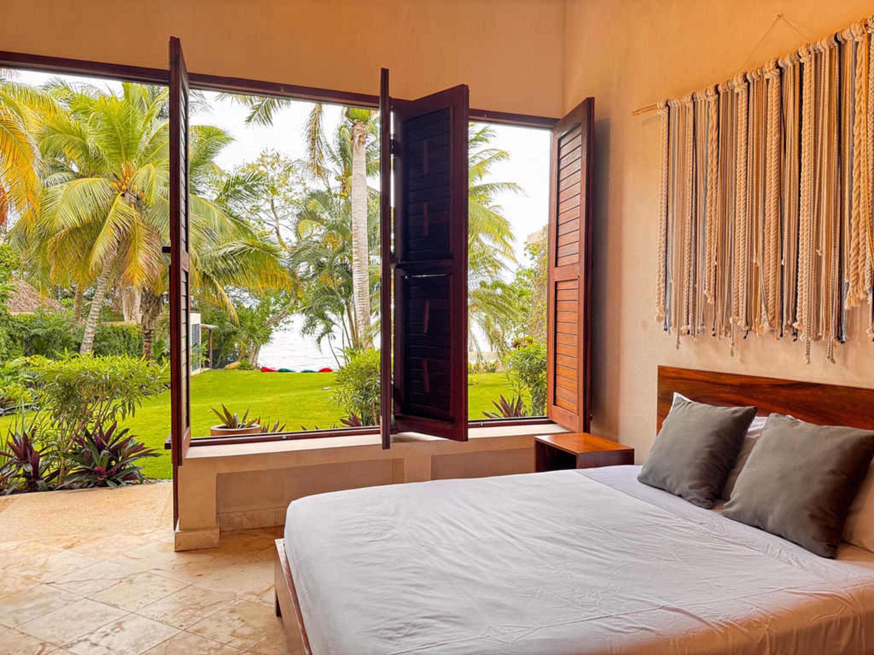Bedroom with a view of palm trees and a grassy yard through open windows.
