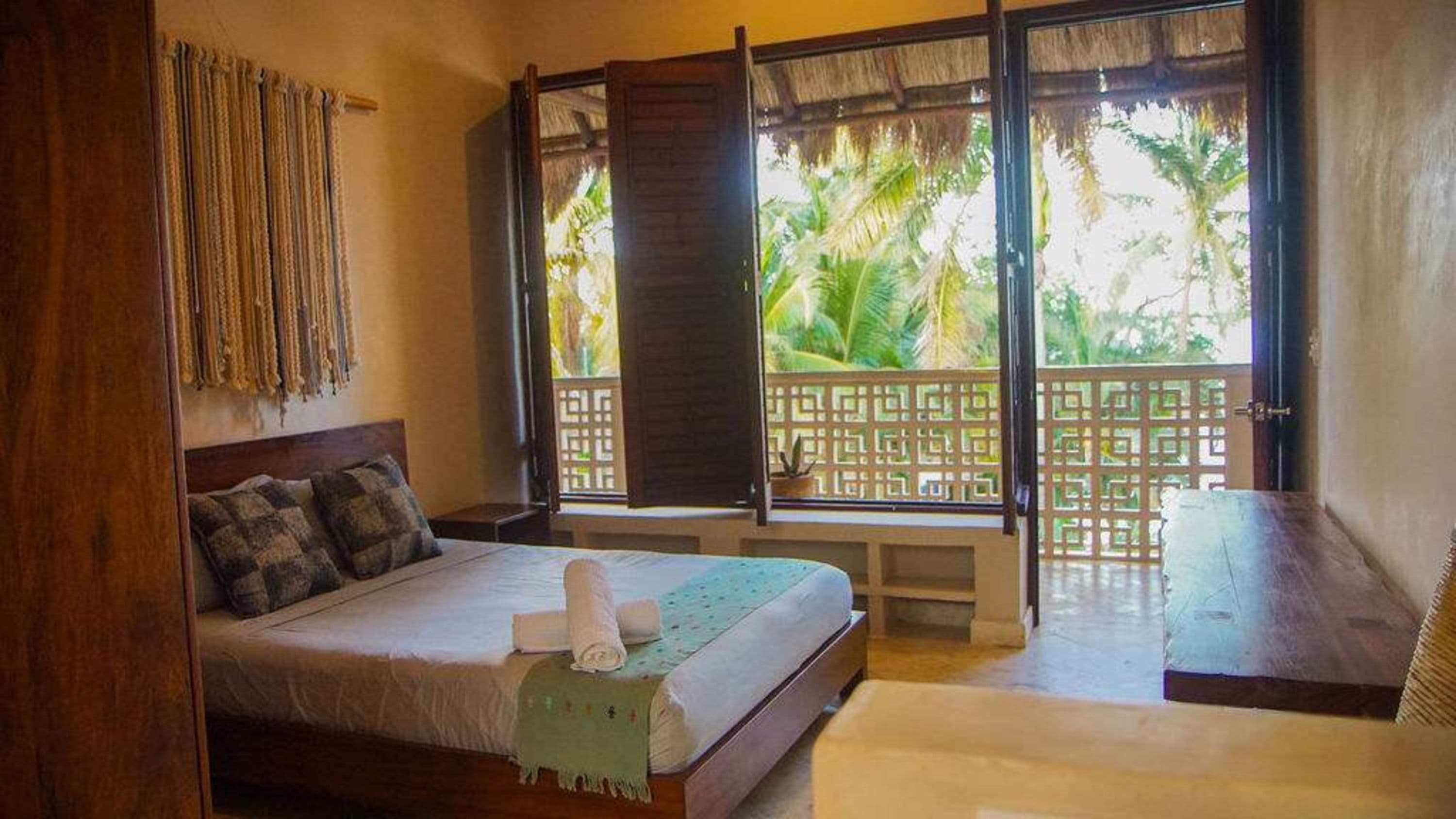A bedroom with a bed, a wooden wardrobe, and a balcony with a view of tropical plants.