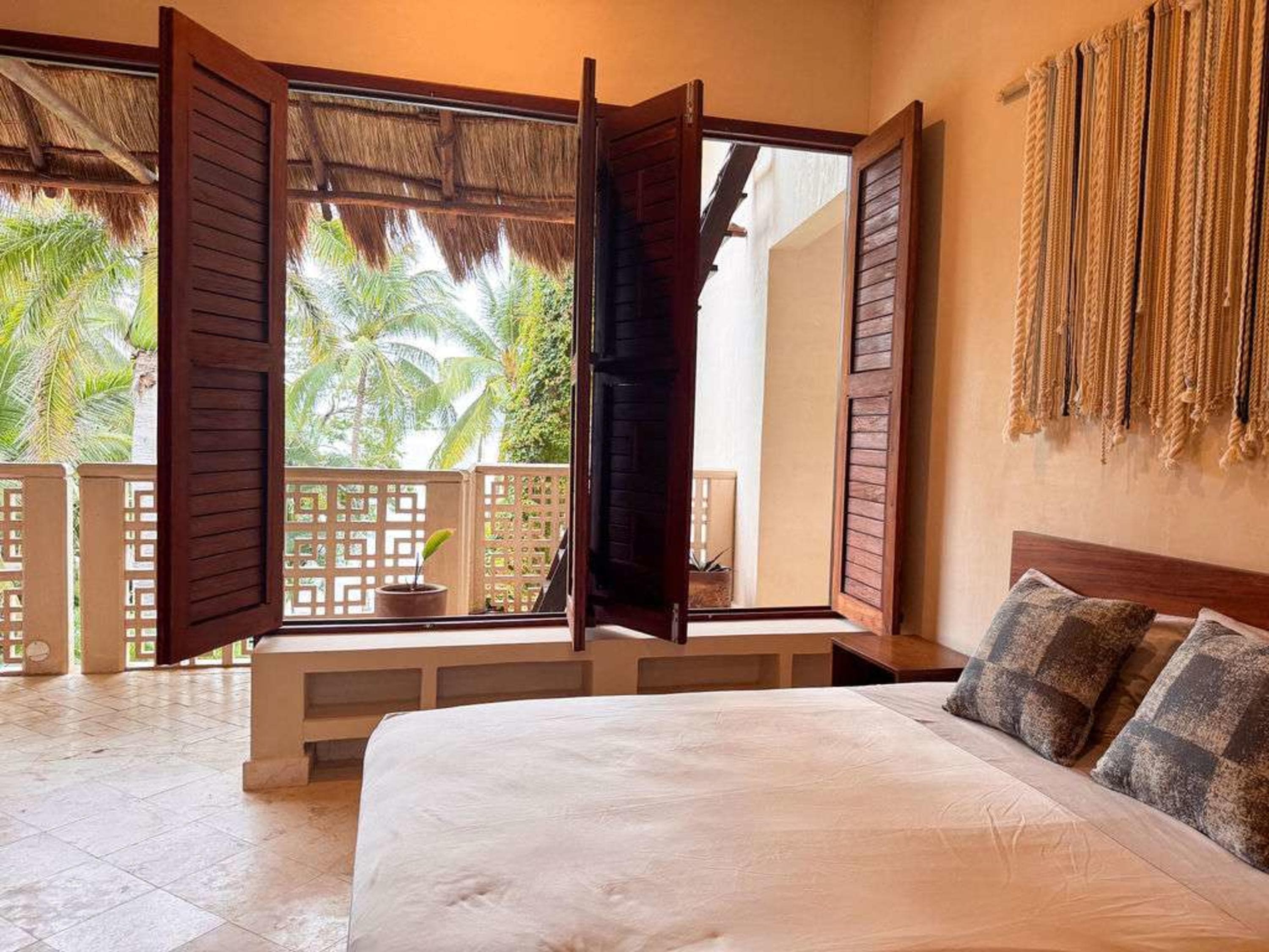 A bedroom with an open wooden shuttered window looking out to palm trees and a balcony.