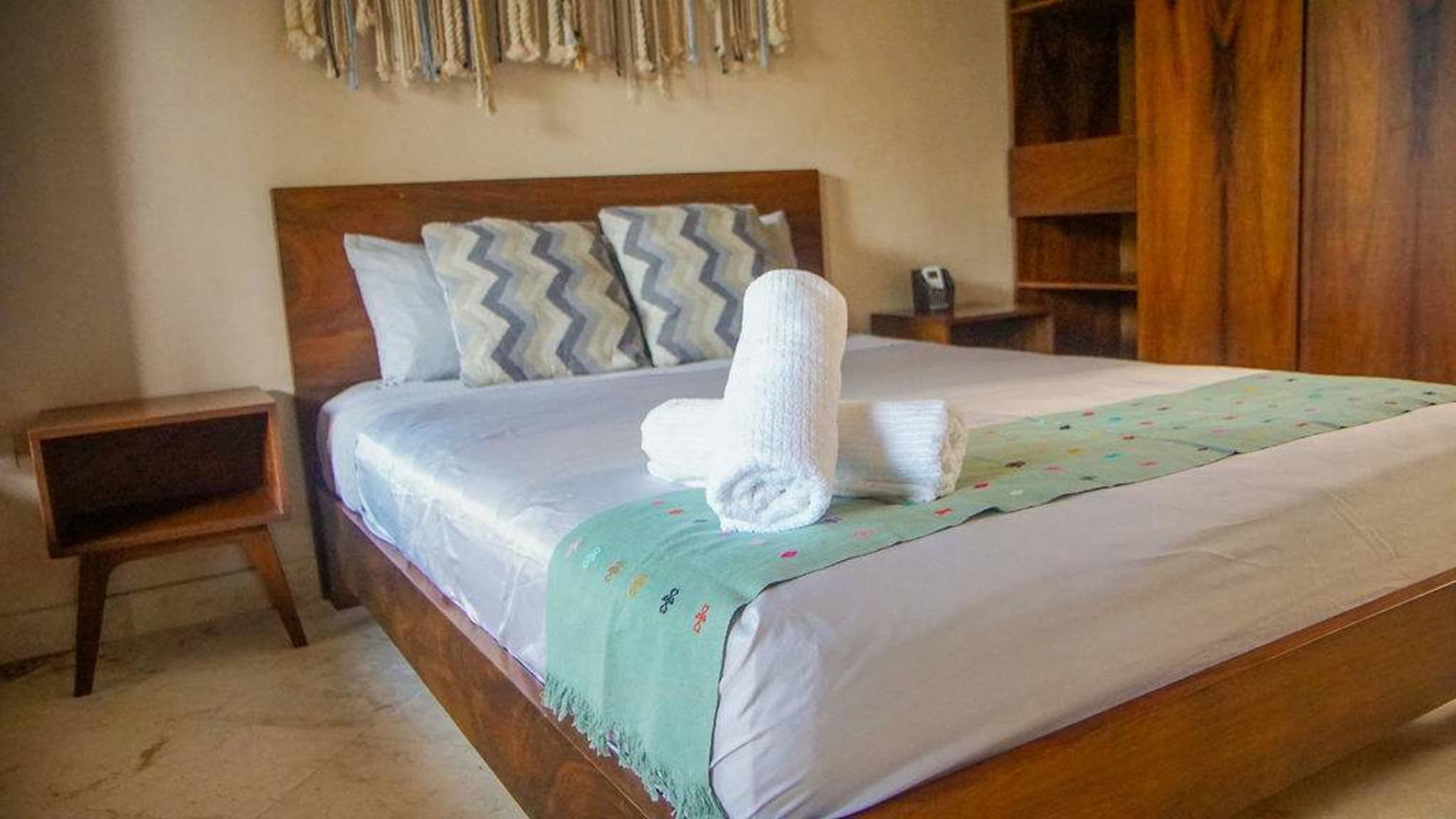 A bed with white linens, two pillows, and two rolled towels sits next to a wooden nightstand.
