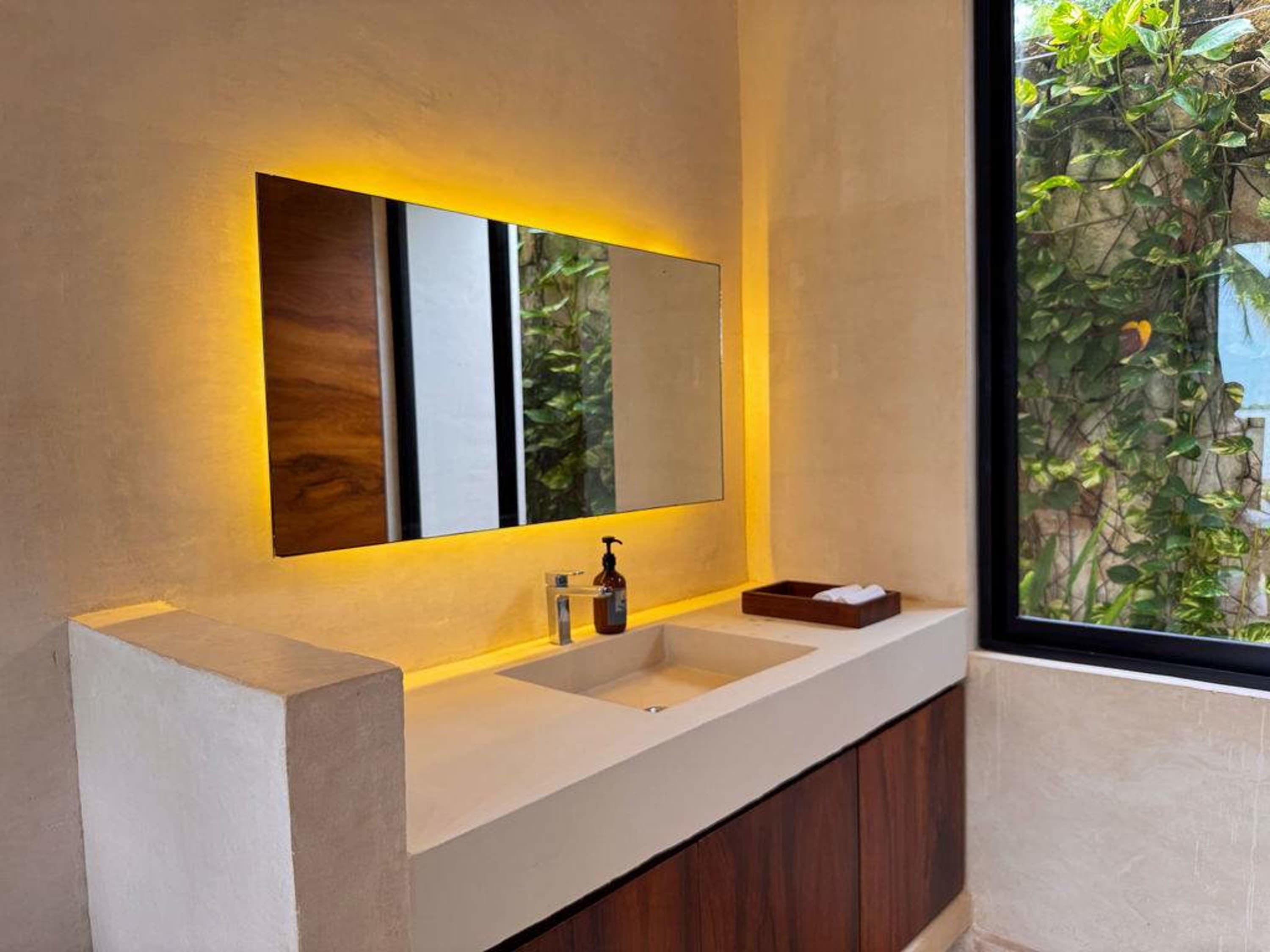 Bathroom vanity with a sink, illuminated mirror, and a view of lush greenery through a window.