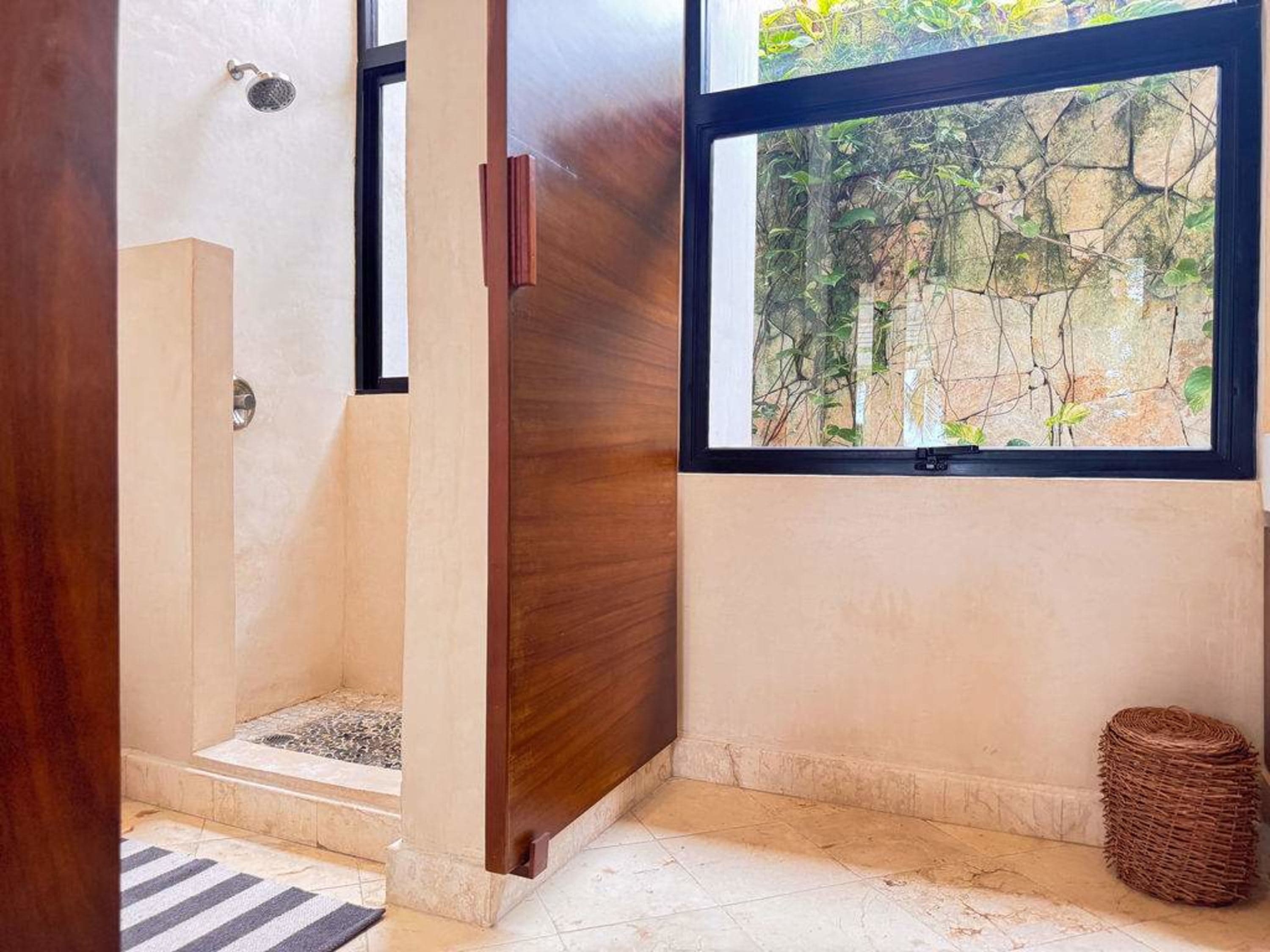 A bathroom with an open shower, a wooden door, and a window looking out at a stone wall and plants.