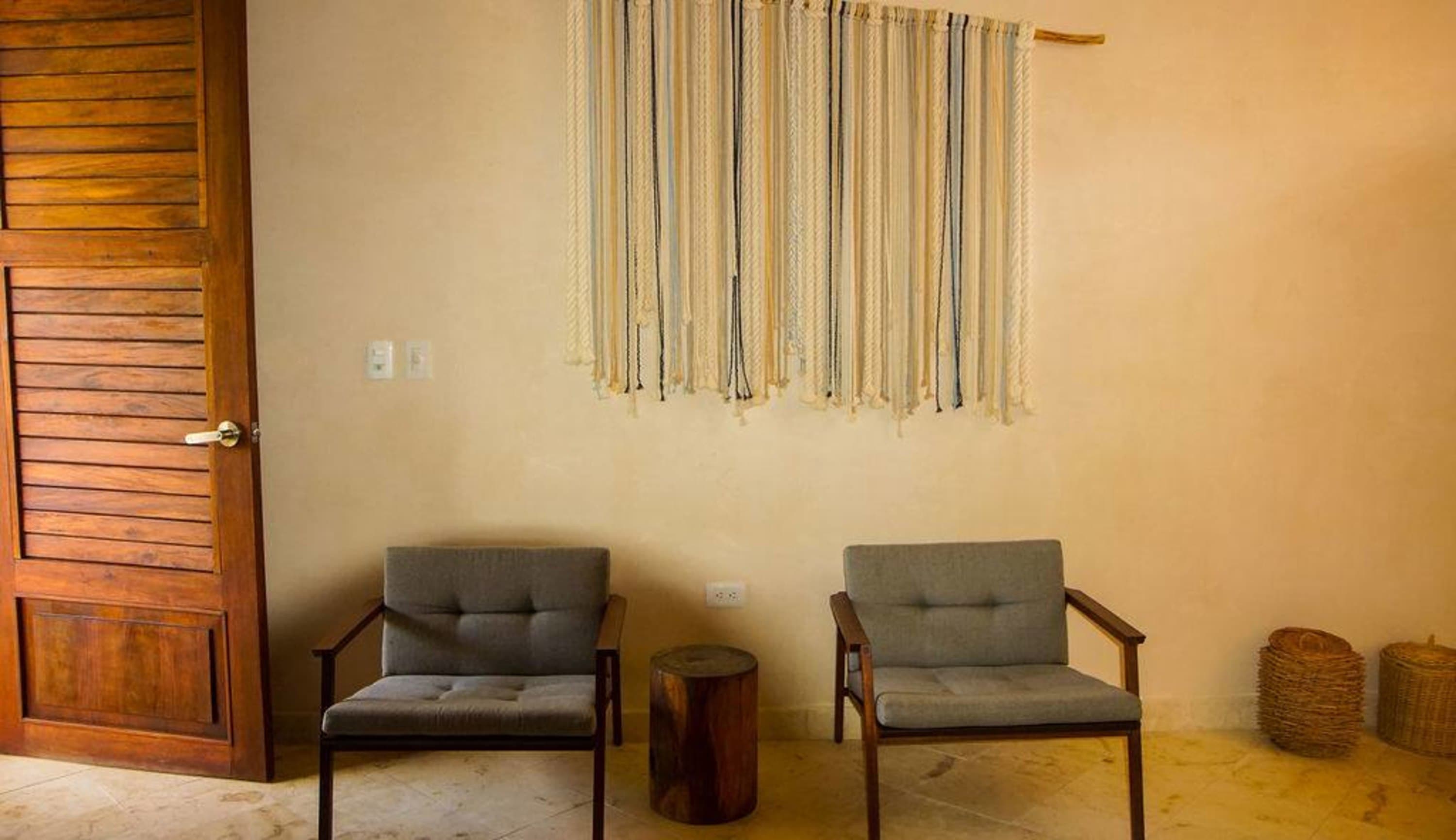 Two grey armchairs flank a wooden stool, with a woven wall hanging above them.