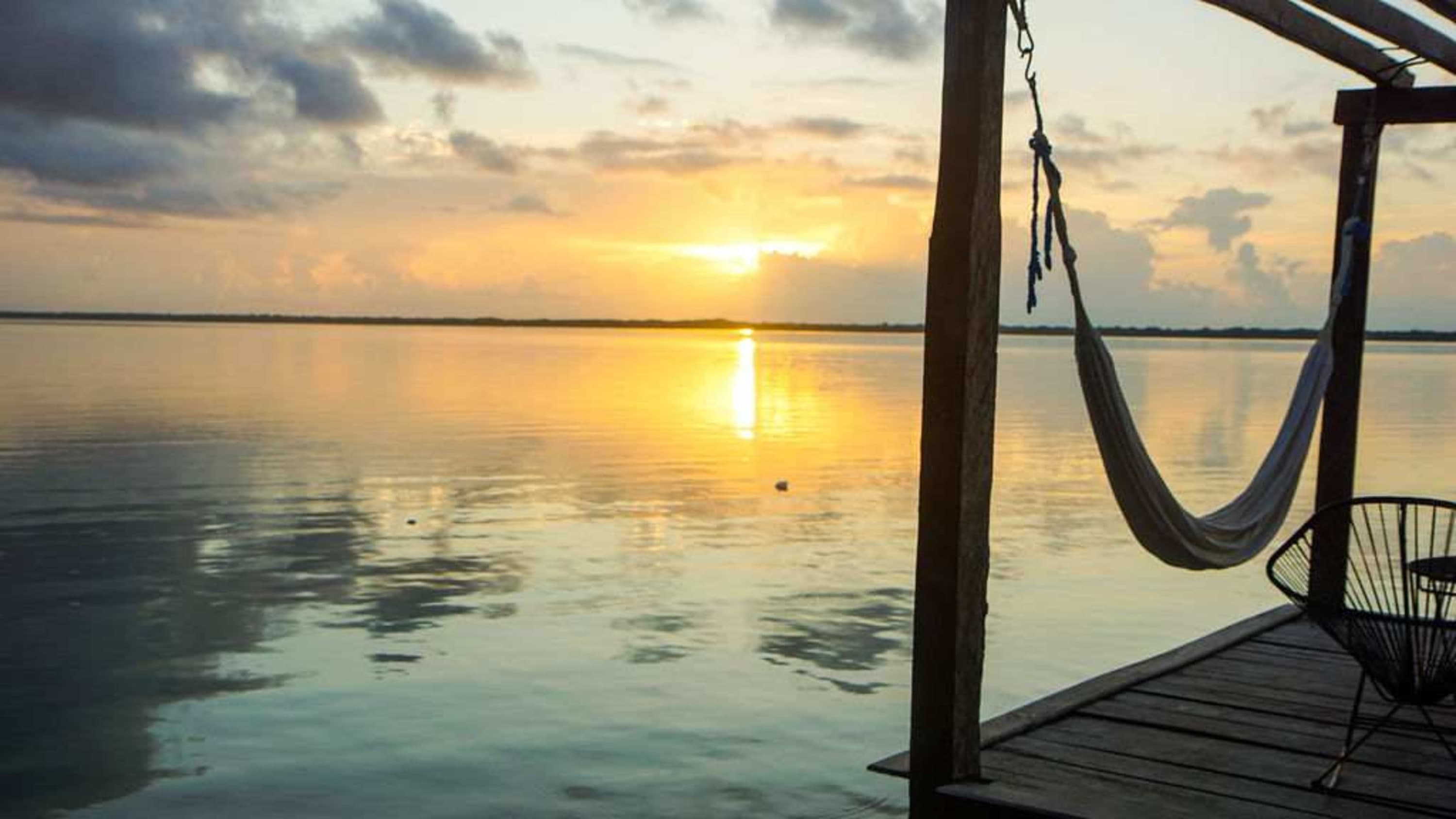A hammock and chair on a dock overlooking a calm lake at sunset.