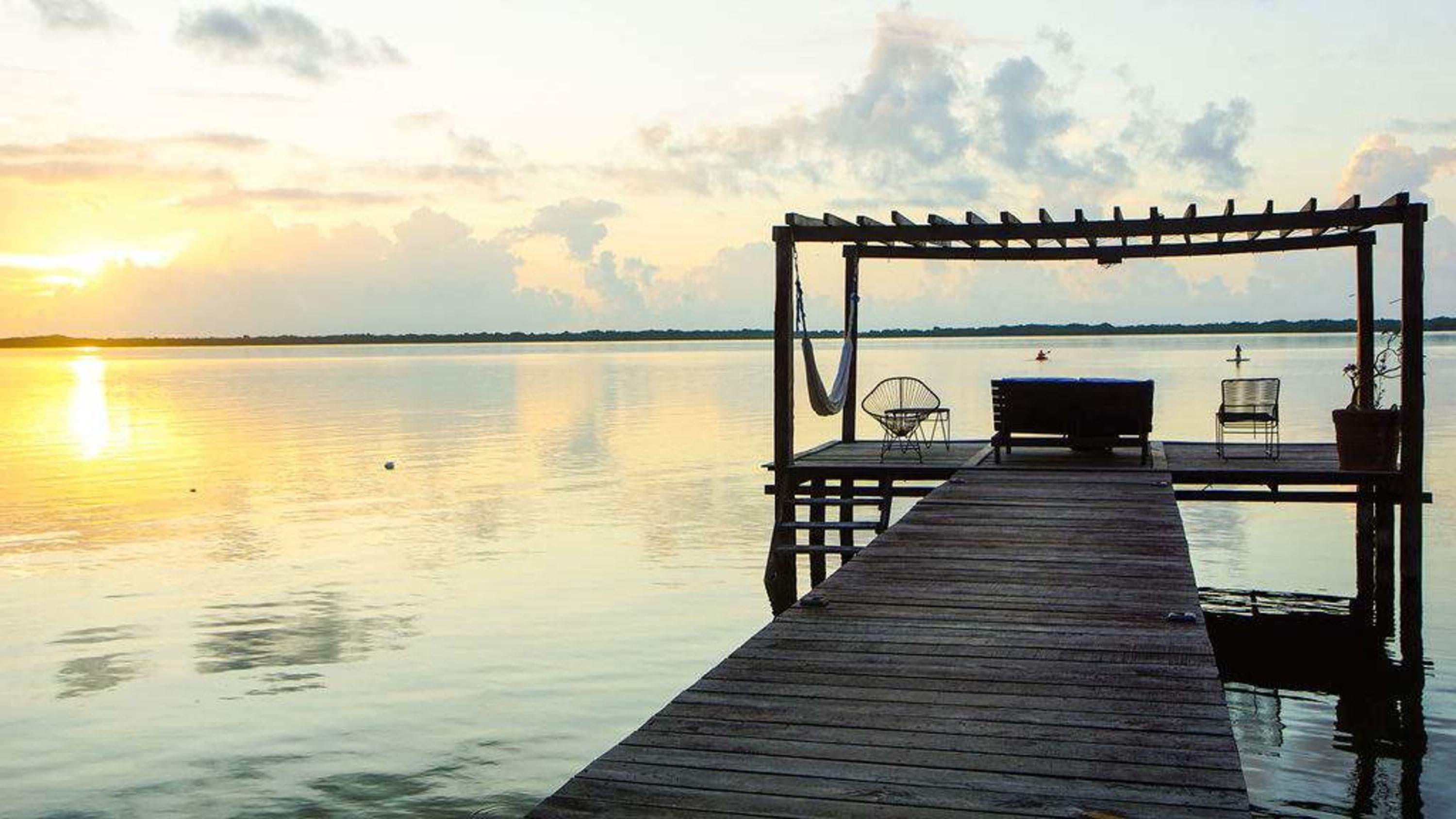A wooden dock with furniture sits over a serene lake at sunset.