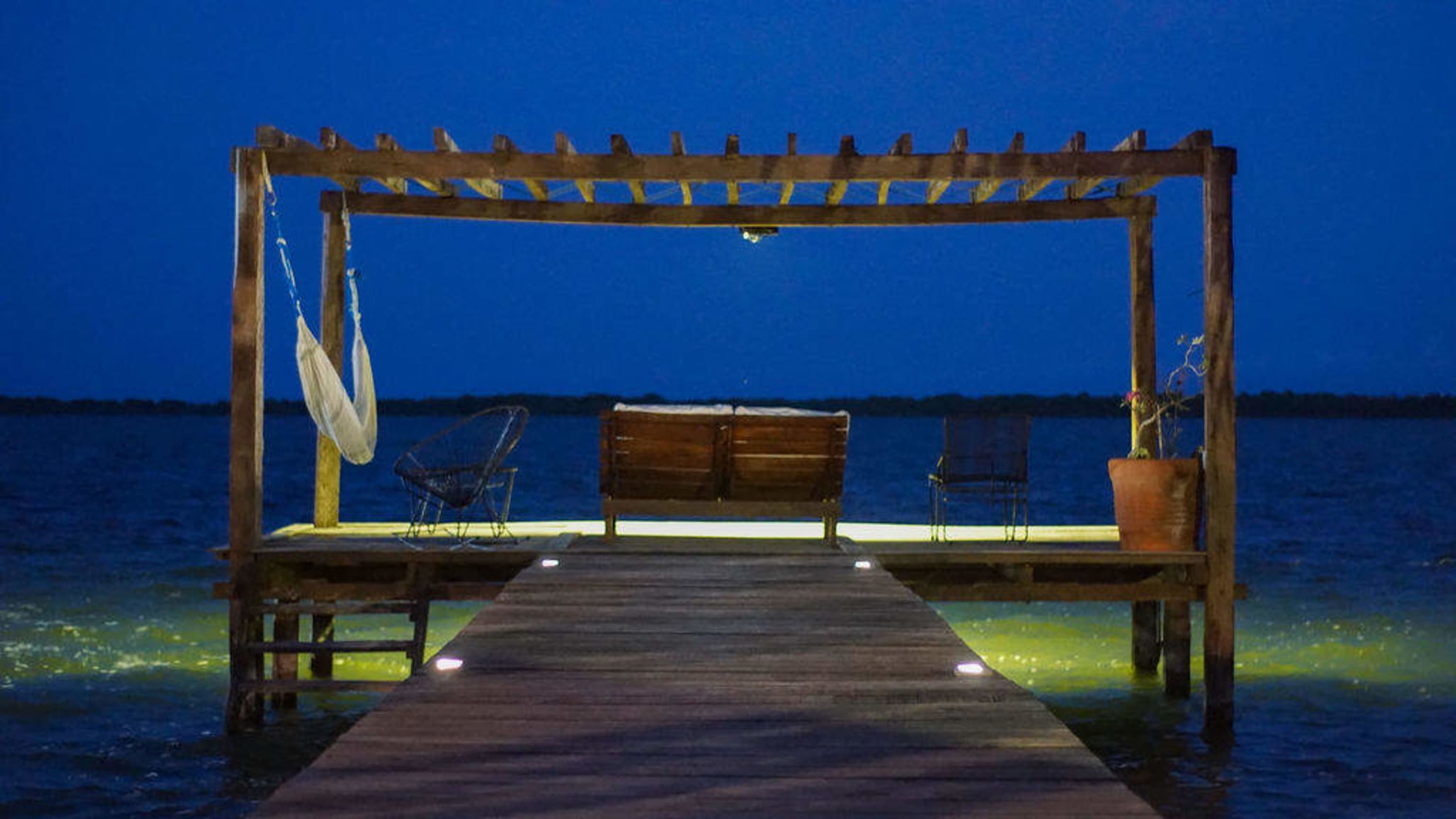 A wooden dock leads to a pier with a pergola, hammock, chairs, and a bench, over a body of water at 