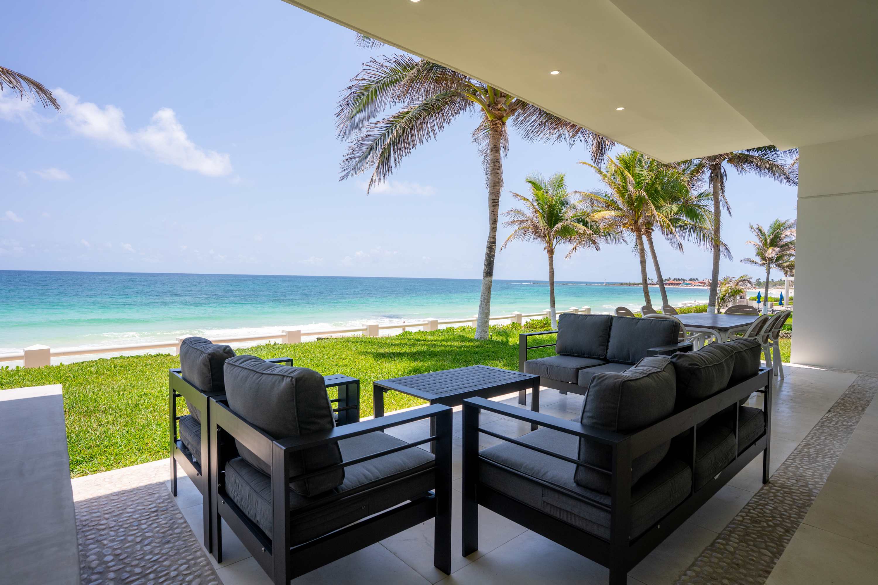 A covered patio with dark gray sofas overlooks a tropical beach with palm trees.