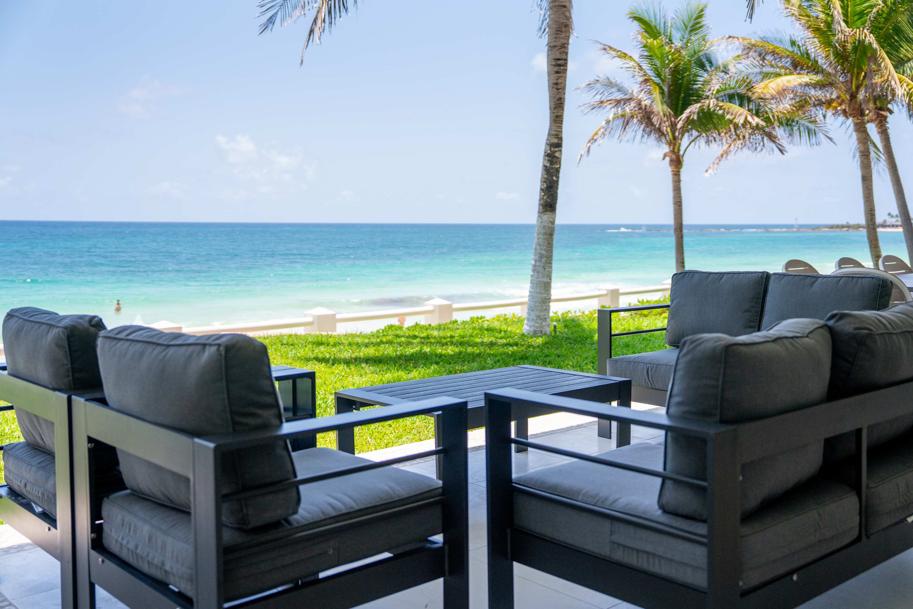 A patio with dark grey seating faces the turquoise ocean, lined with palm trees.