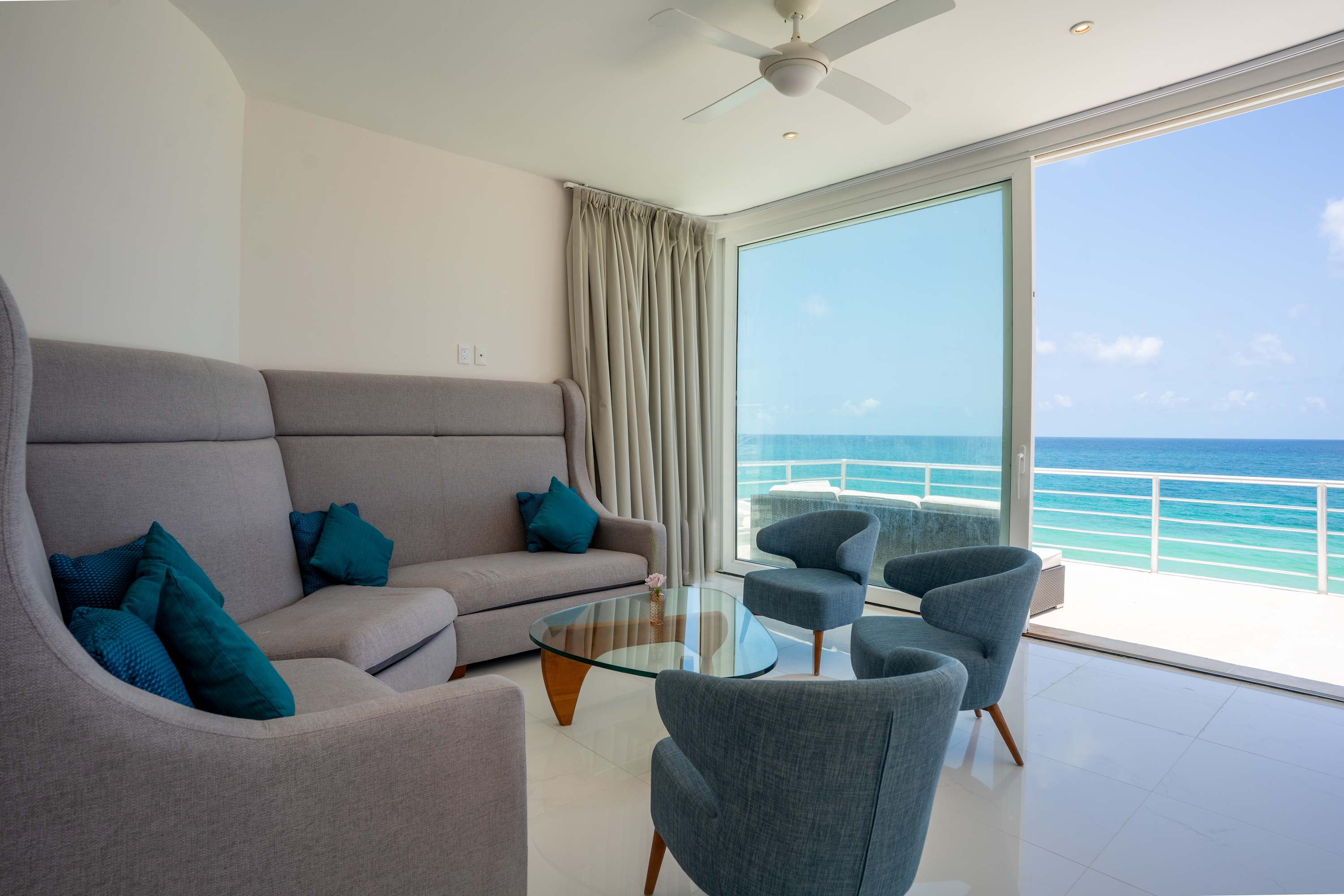 A living room with a sofa, armchairs, and coffee table faces a balcony with a view of the ocean.