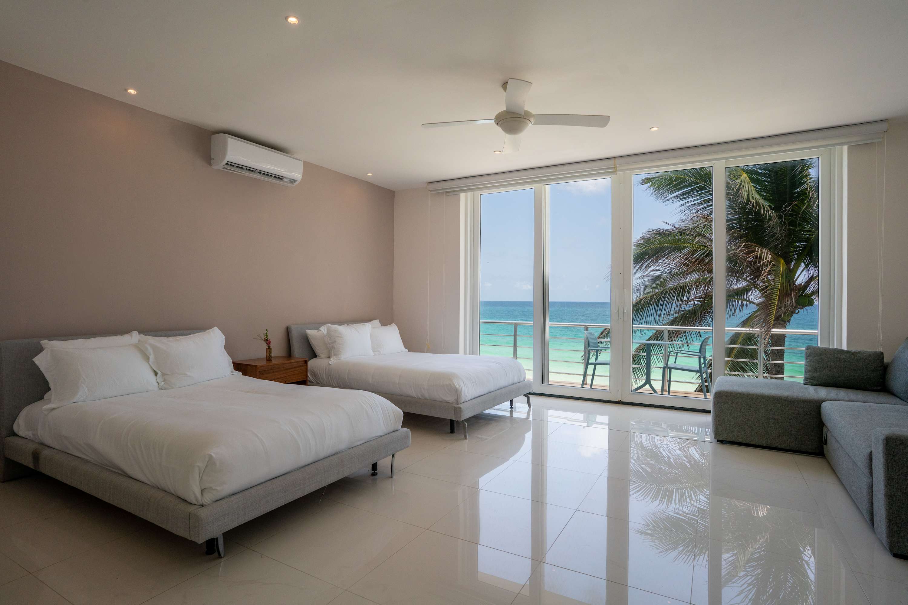 A modern bedroom with two beds and a gray sofa facing the ocean view.