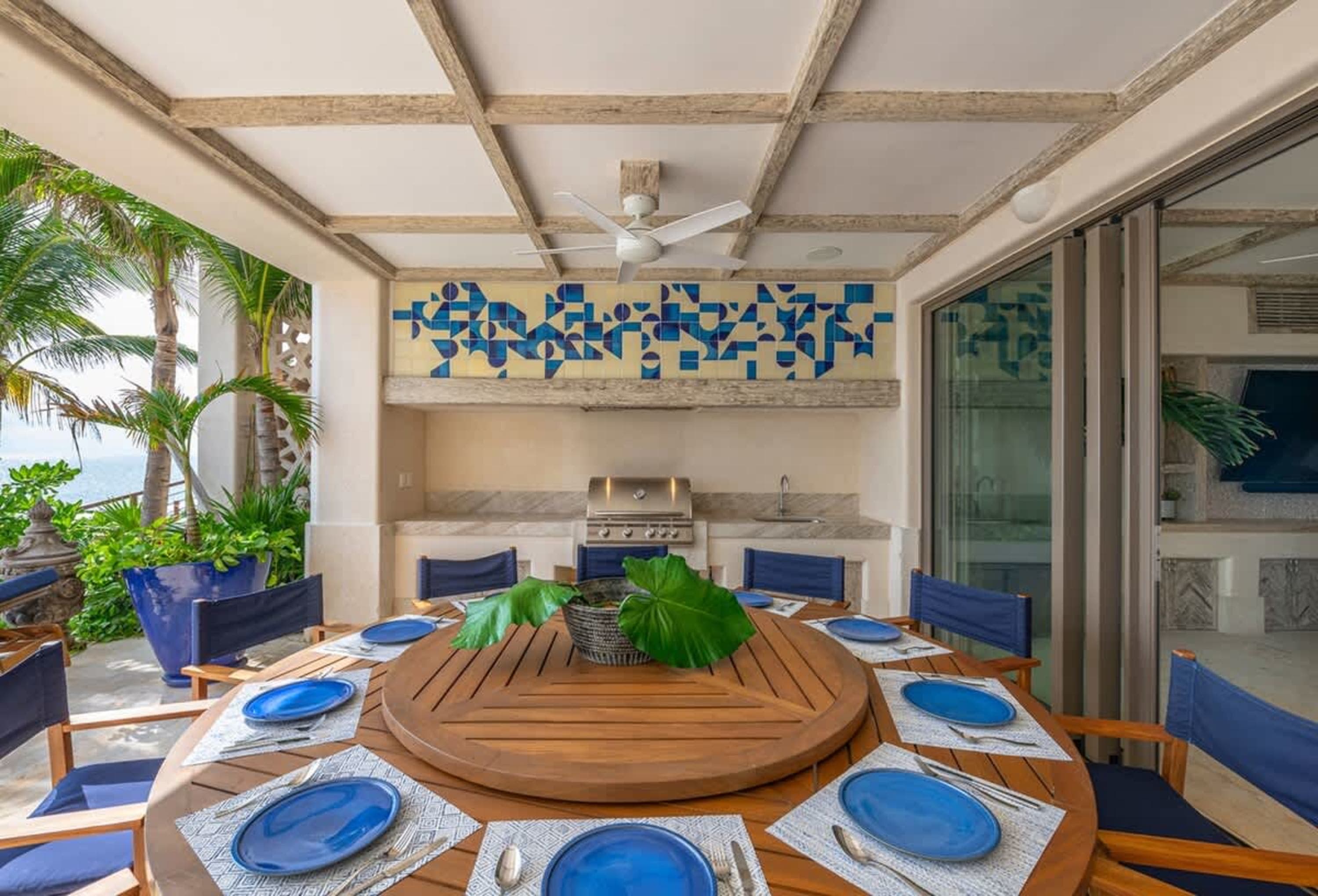 A covered patio with a dining table set for a meal, a grill and sink area, and palm trees.