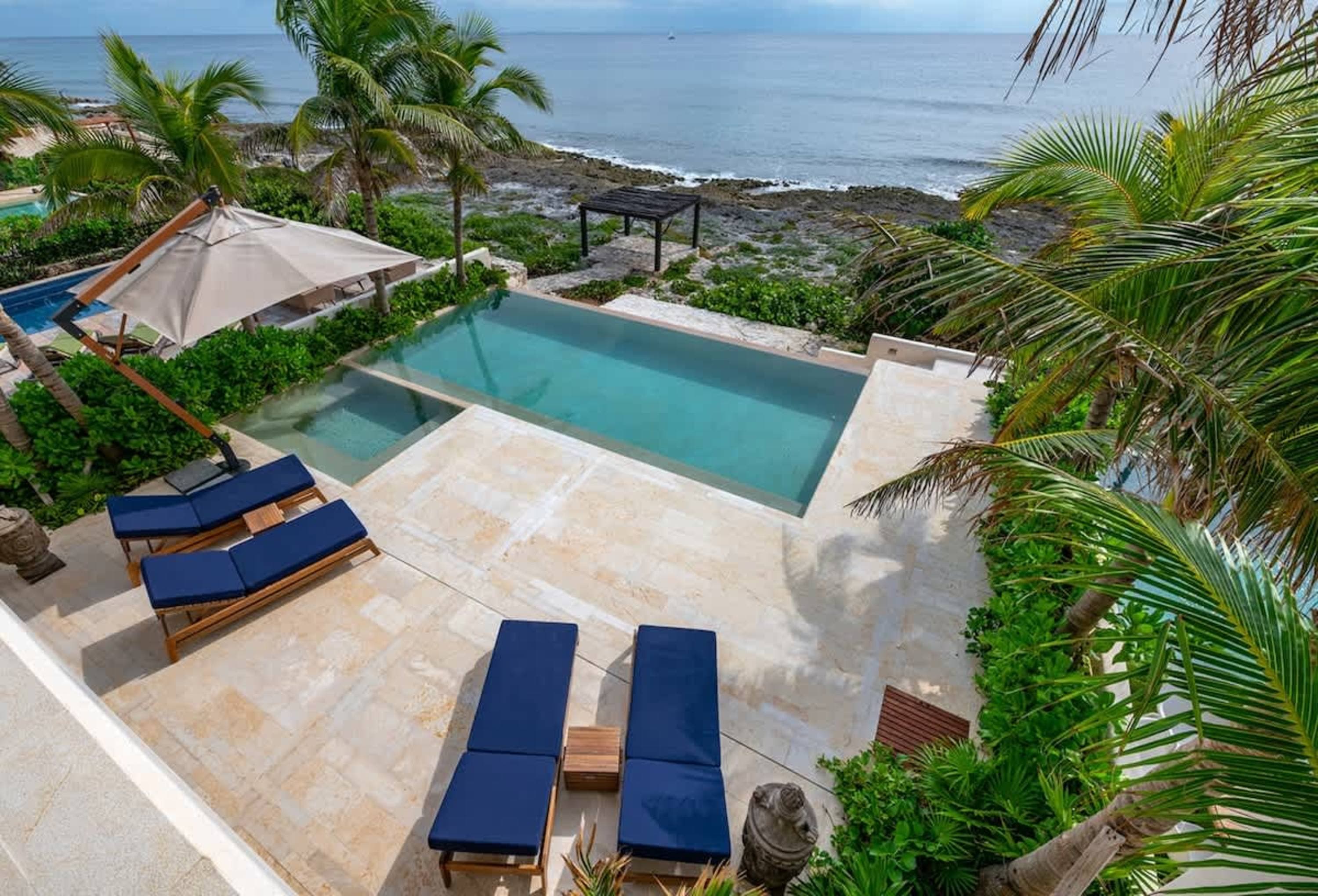 A luxurious infinity pool and lounge chairs overlook the ocean, surrounded by palm trees and lush gr