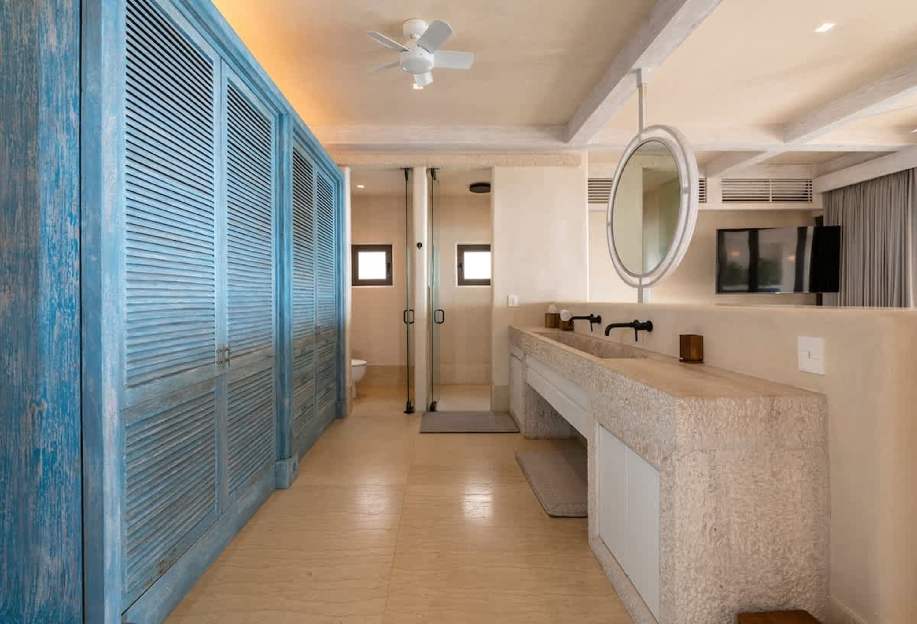 A spacious bathroom with blue louvered cabinets, a double vanity with stone countertop, and a glass-