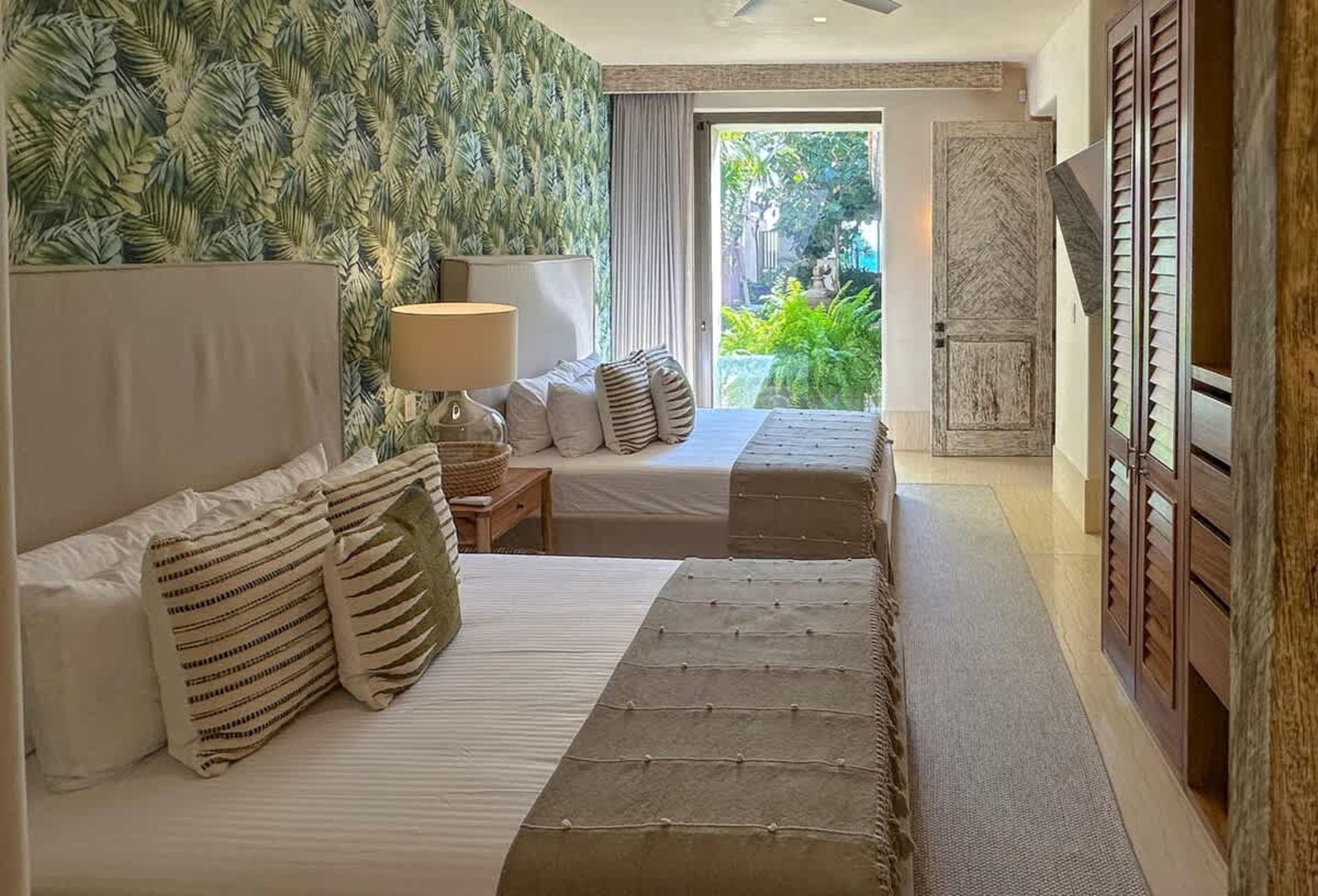 A hotel room with two beds, a tropical wallpaper, and a view of greenery.