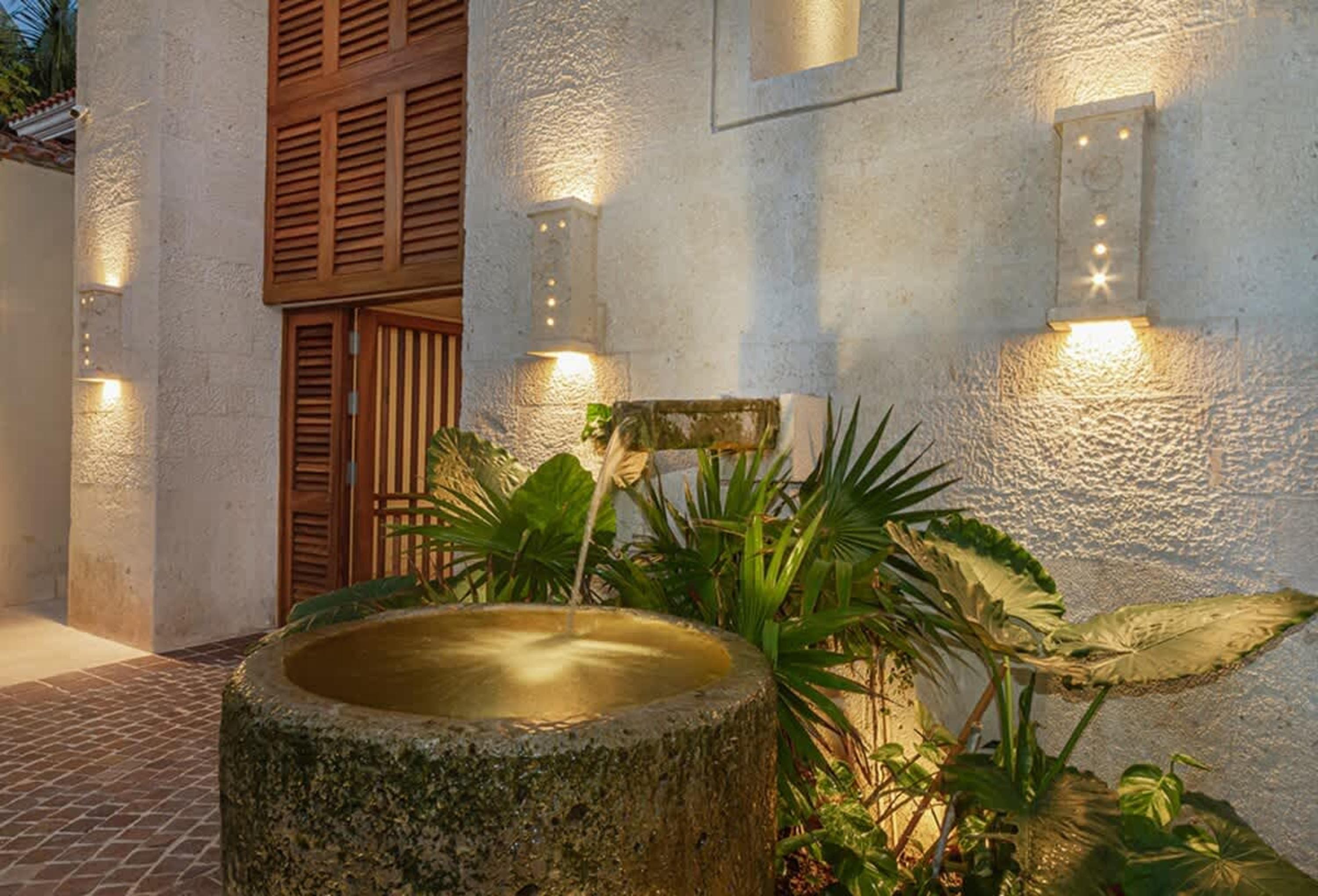 Exterior view of a stone building with wooden doors, a fountain, lush plants, and illuminated wall s