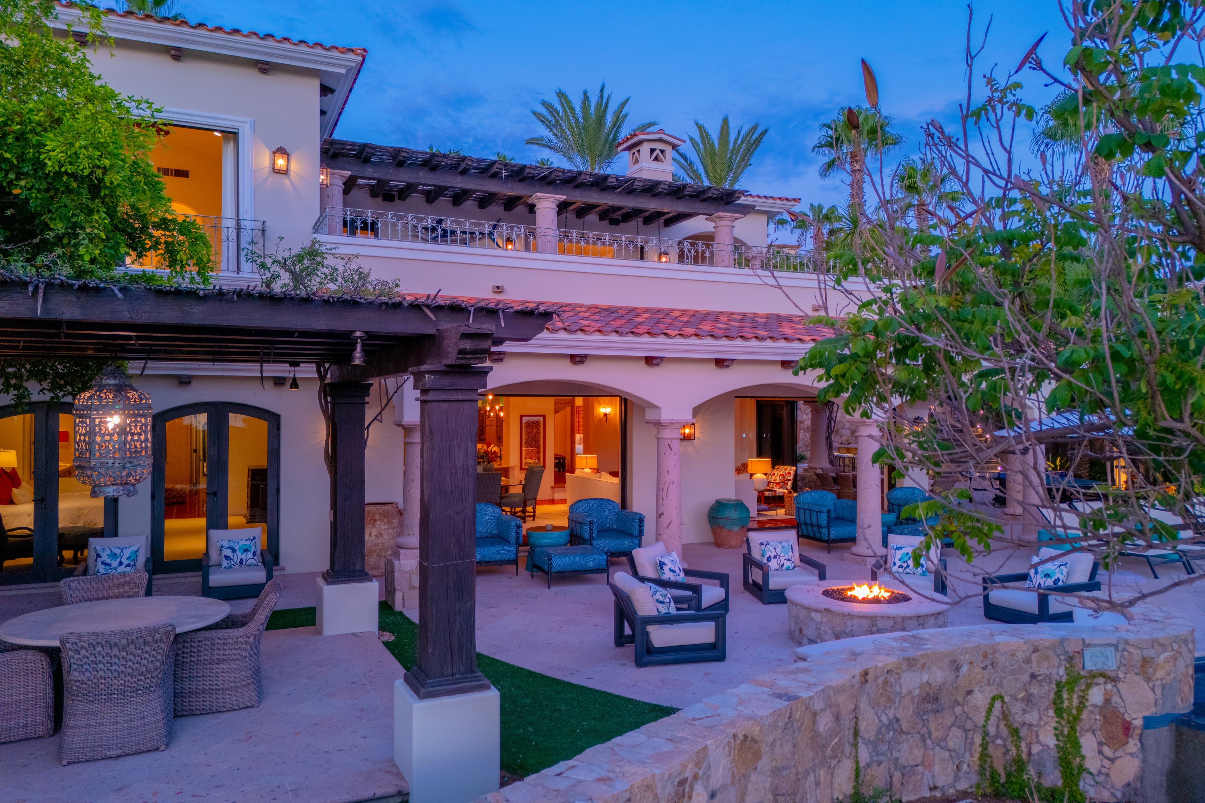 A stunning Mediterranean-style villa photographed at twilight, featuring multi-level architecture with red-tile roofing, arched doorways, and lush tropical landscaping. The property's elegant outdoor living spaces are softly illuminated against the evening