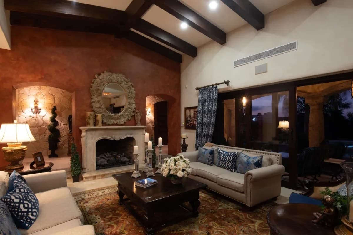 Unwind in your elegant living room with vaulted ceilings, exposed beams, a charming fireplace, and comfortable seating. Large windows frame peaceful evening views, creating the perfect retreat.
