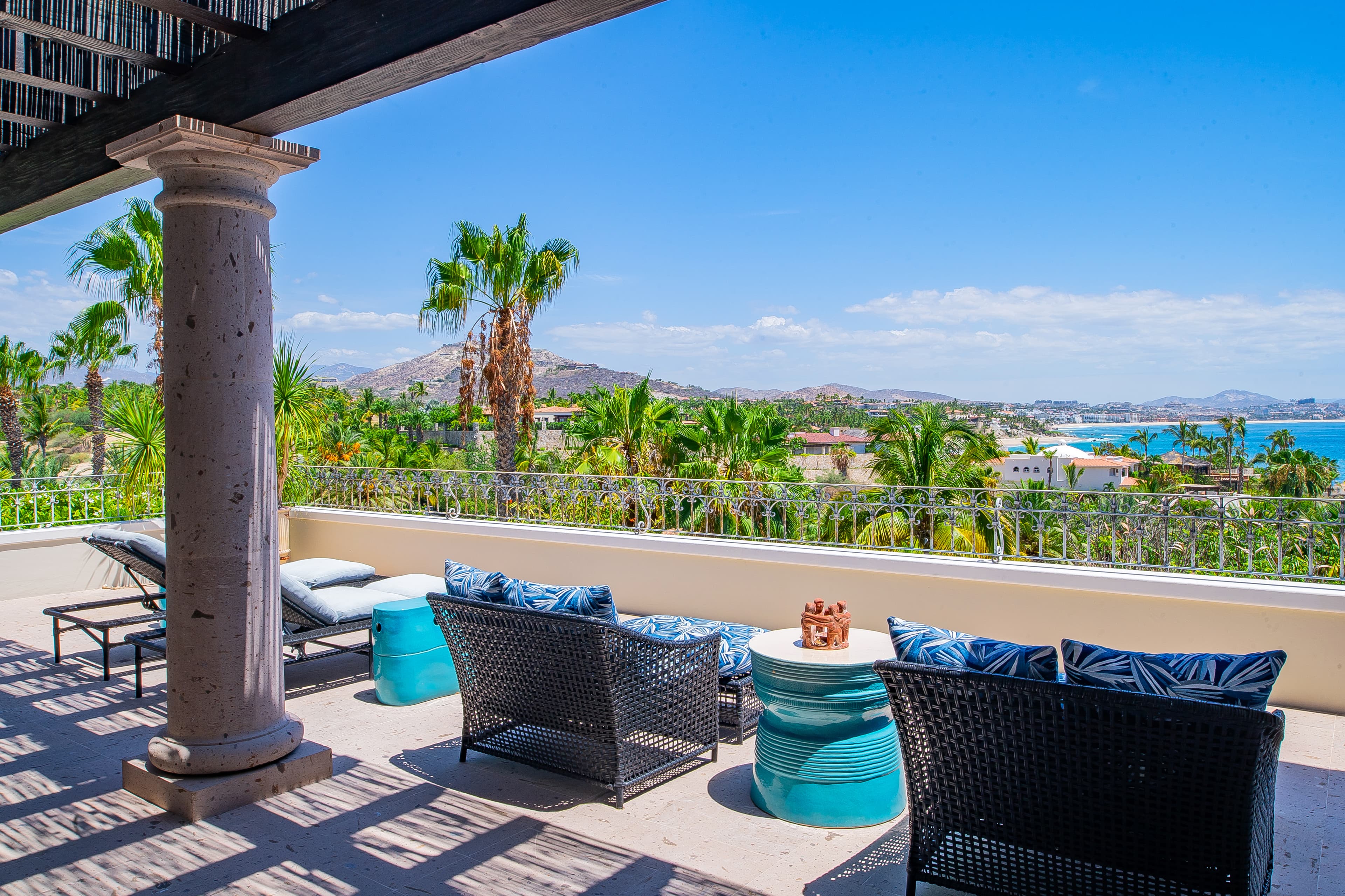 Relax on your private terrace with turquoise seating under shaded columns while taking in panoramic views of swaying palm trees, hillside villas, and sparkling turquoise sea stretching toward distant mountains.