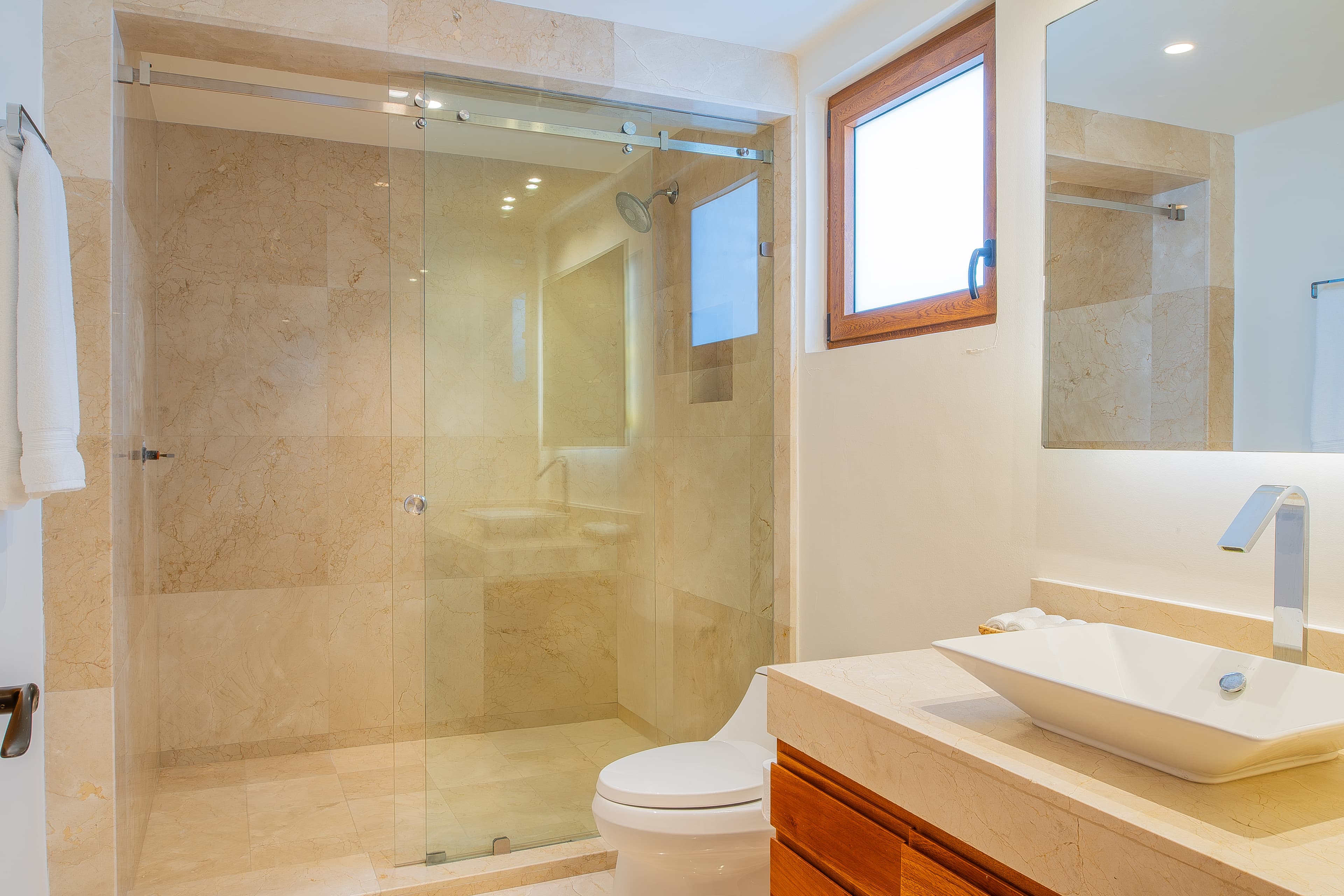 Bathroom 4 · Step into your spa-like bathroom featuring warm travertine tile, a sleek modern vanity, and a spacious glass-enclosed shower. Natural light pours in through the wood-framed window, setting the perfect mood.