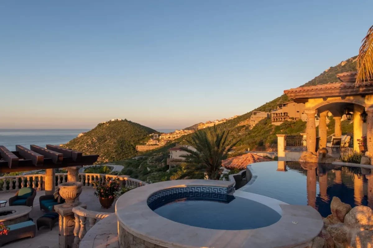 Hillside villa features infinity pool with panoramic ocean and mountain views at golden hour, showcasing Mediterranean-style architecture and lush coastal landscape.