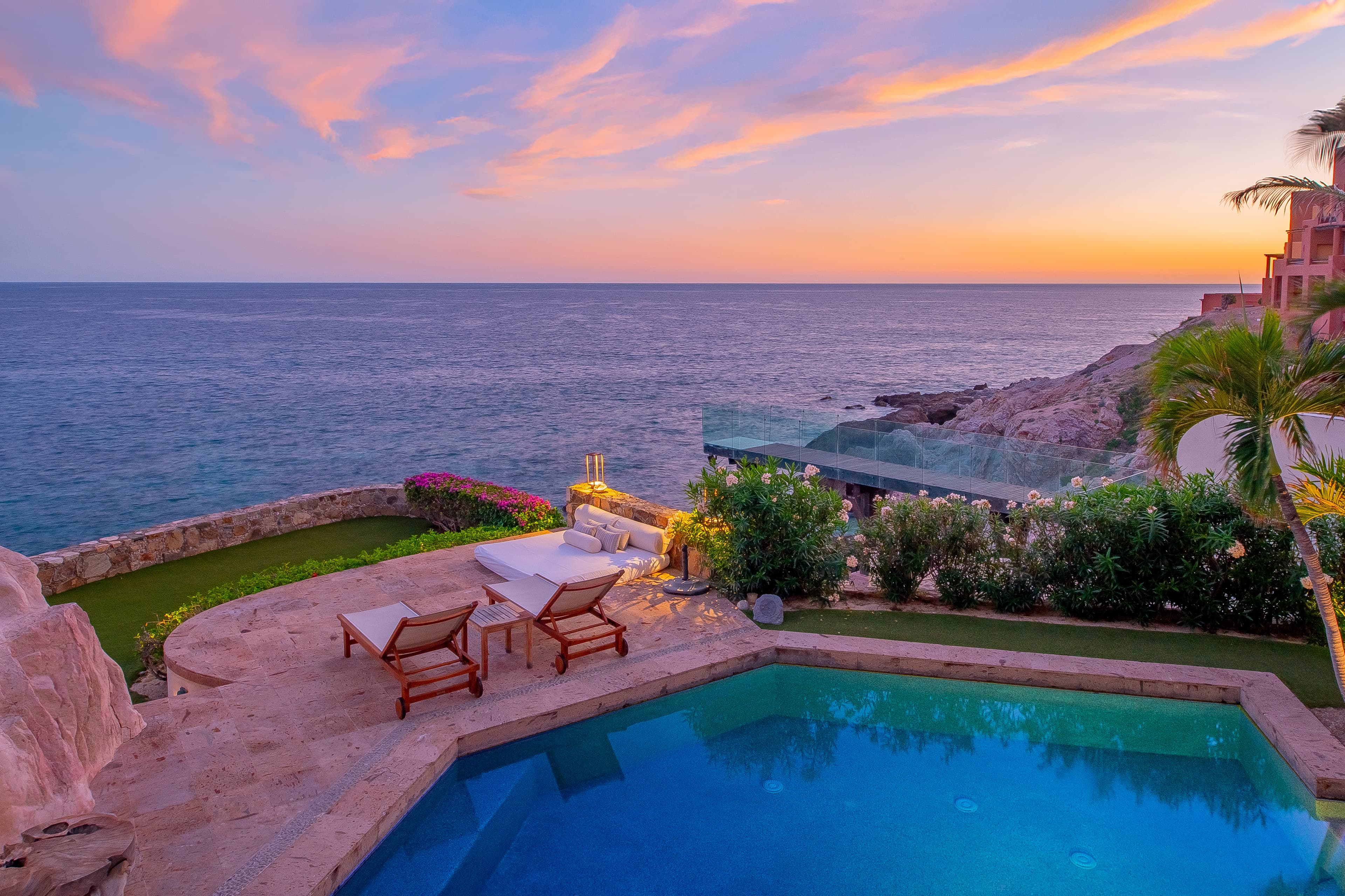 Stunning seaside villa with private pool overlooking tranquil ocean waters at sunset, surrounded by lush tropical gardens and terraced lawns cascading toward the rocky shoreline.