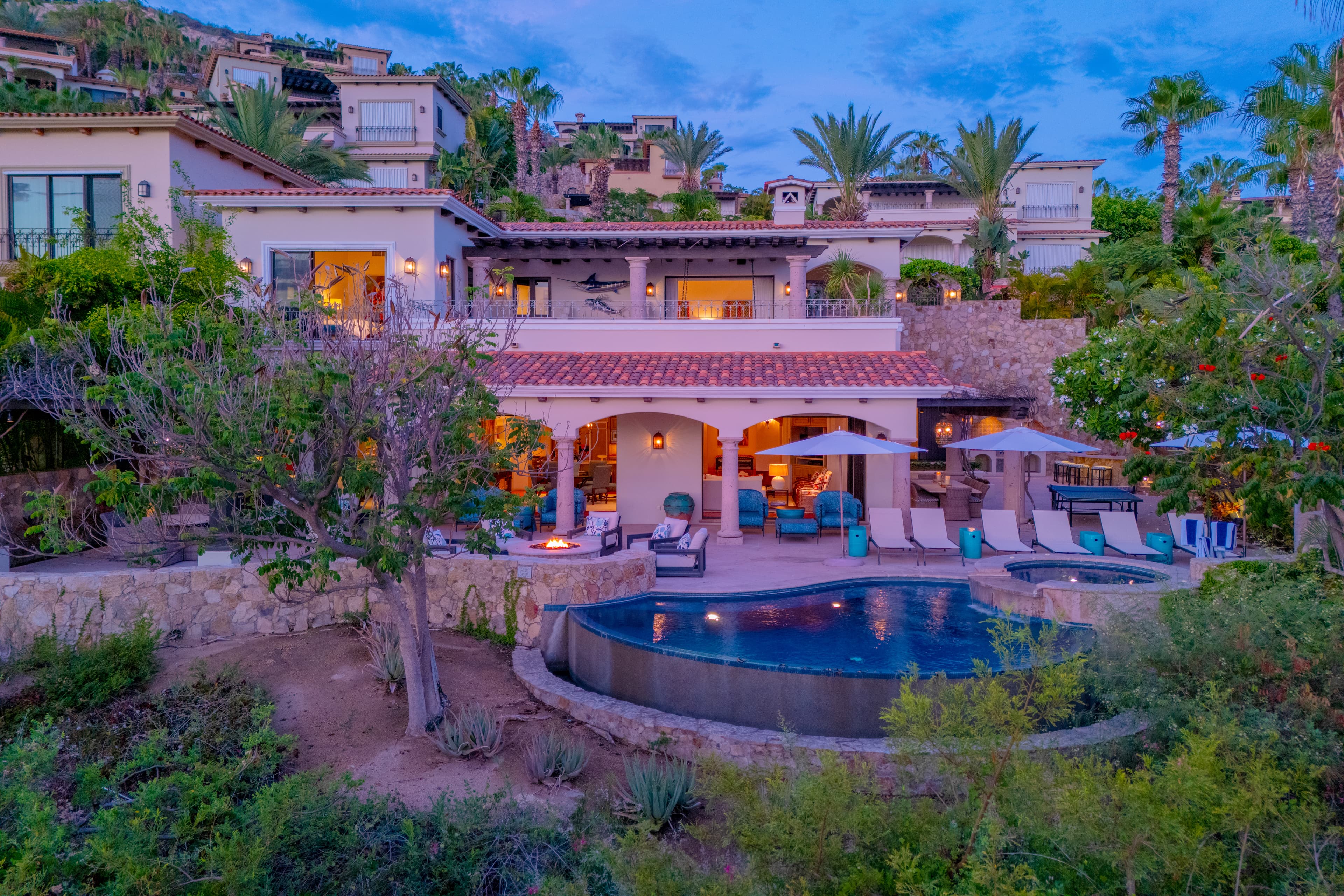 Stunning hillside villa nestled among palms and lush gardens, featuring private pool, multiple terraces, and dramatic views at twilight.