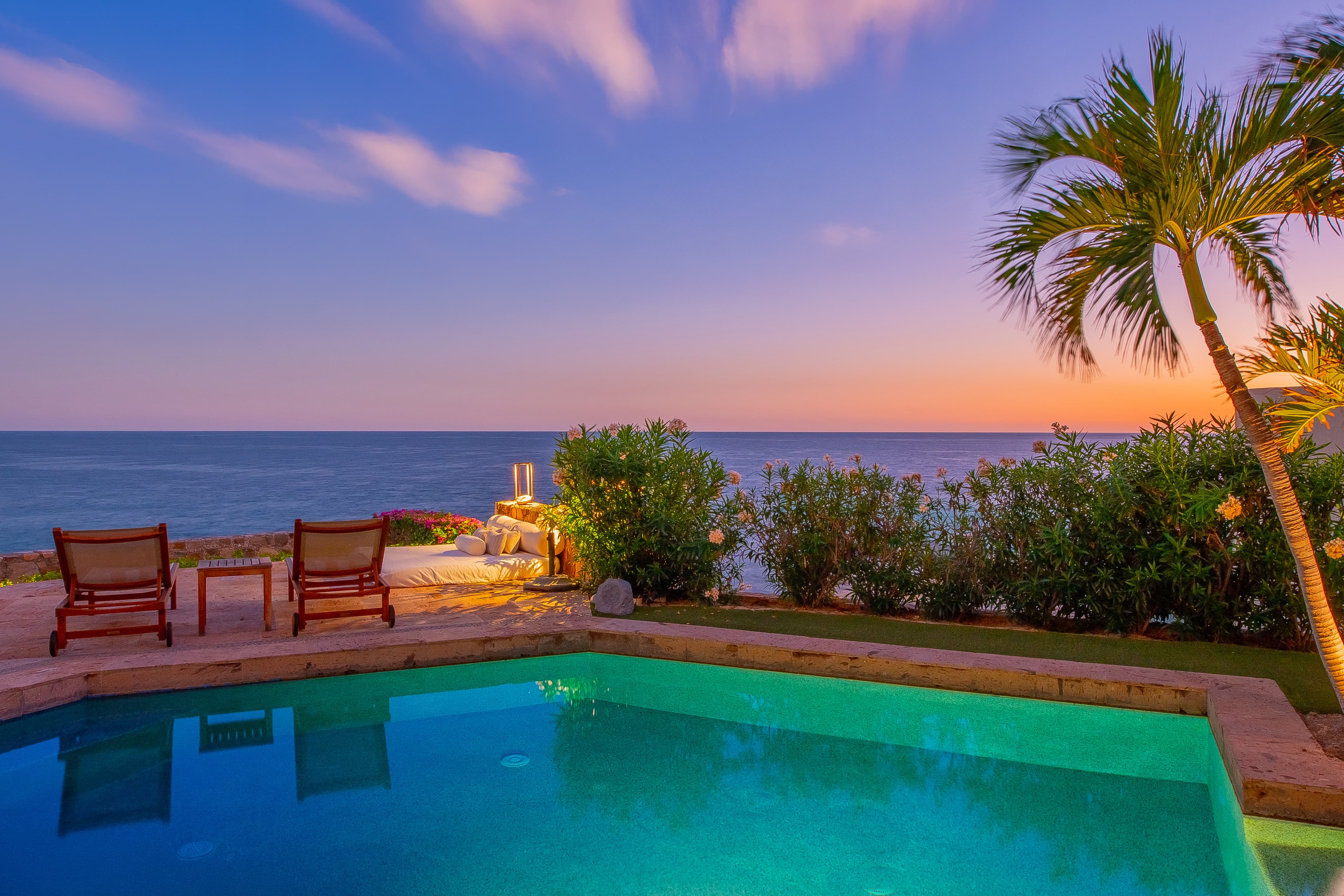 A tranquil oceanfront pool awaits as the sky transforms into soft pastels at dusk, with palm trees swaying and lounge chairs inviting relaxation by the water's edge.