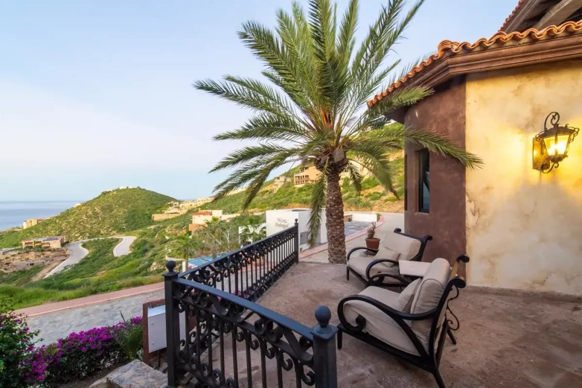 A hillside terrace overlooking the Sea of Cortez, featuring Mediterranean-style architecture with terra cotta roof tiles, decorative ironwork, and lush tropical landscaping in a serene coastal setting.