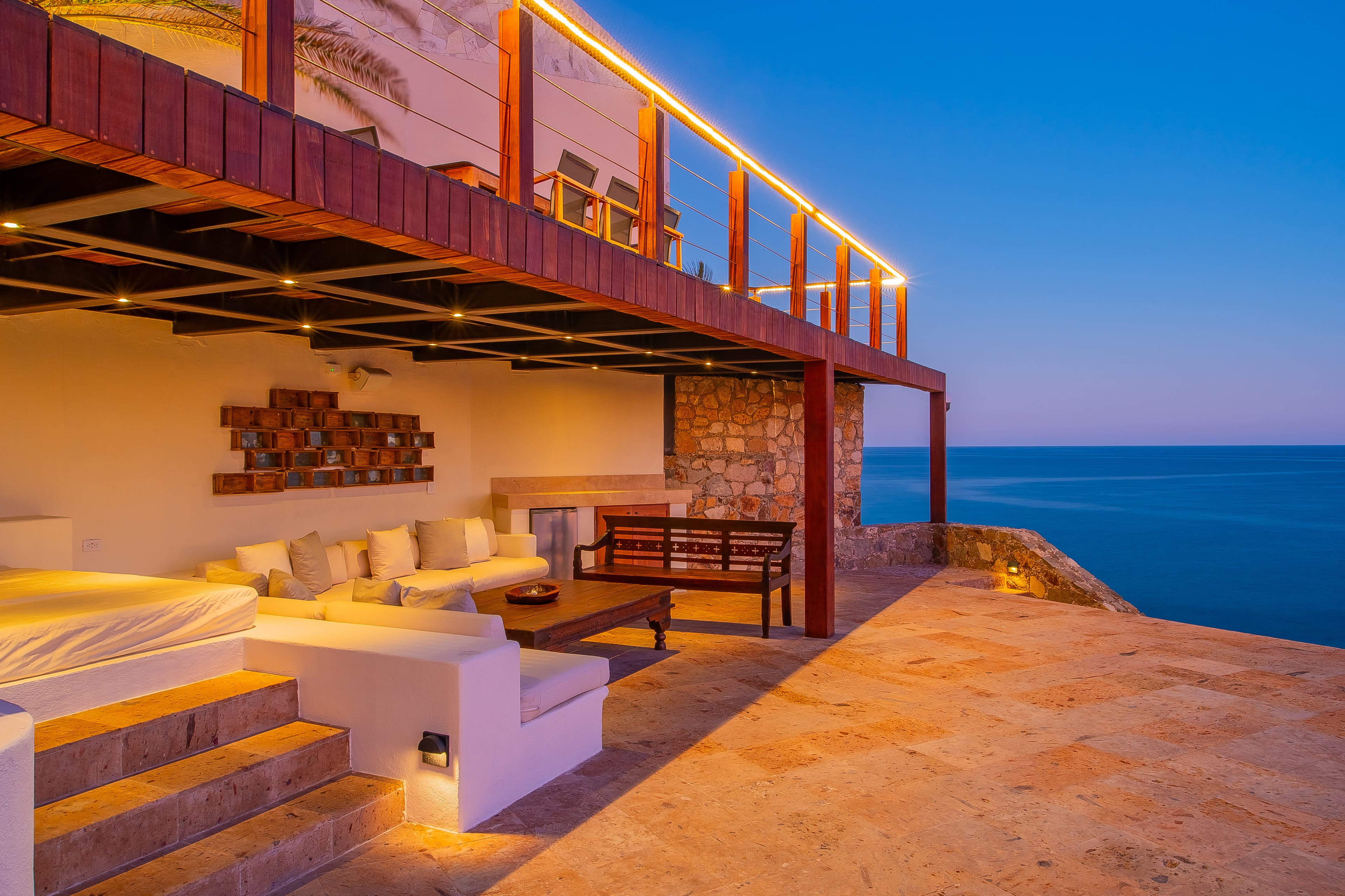 Stunning oceanfront villa showcasing multi-level architecture, elegant stone and wood design, ambient lighting at dusk, and an expansive terrace with breathtaking sea views stretching to the horizon.