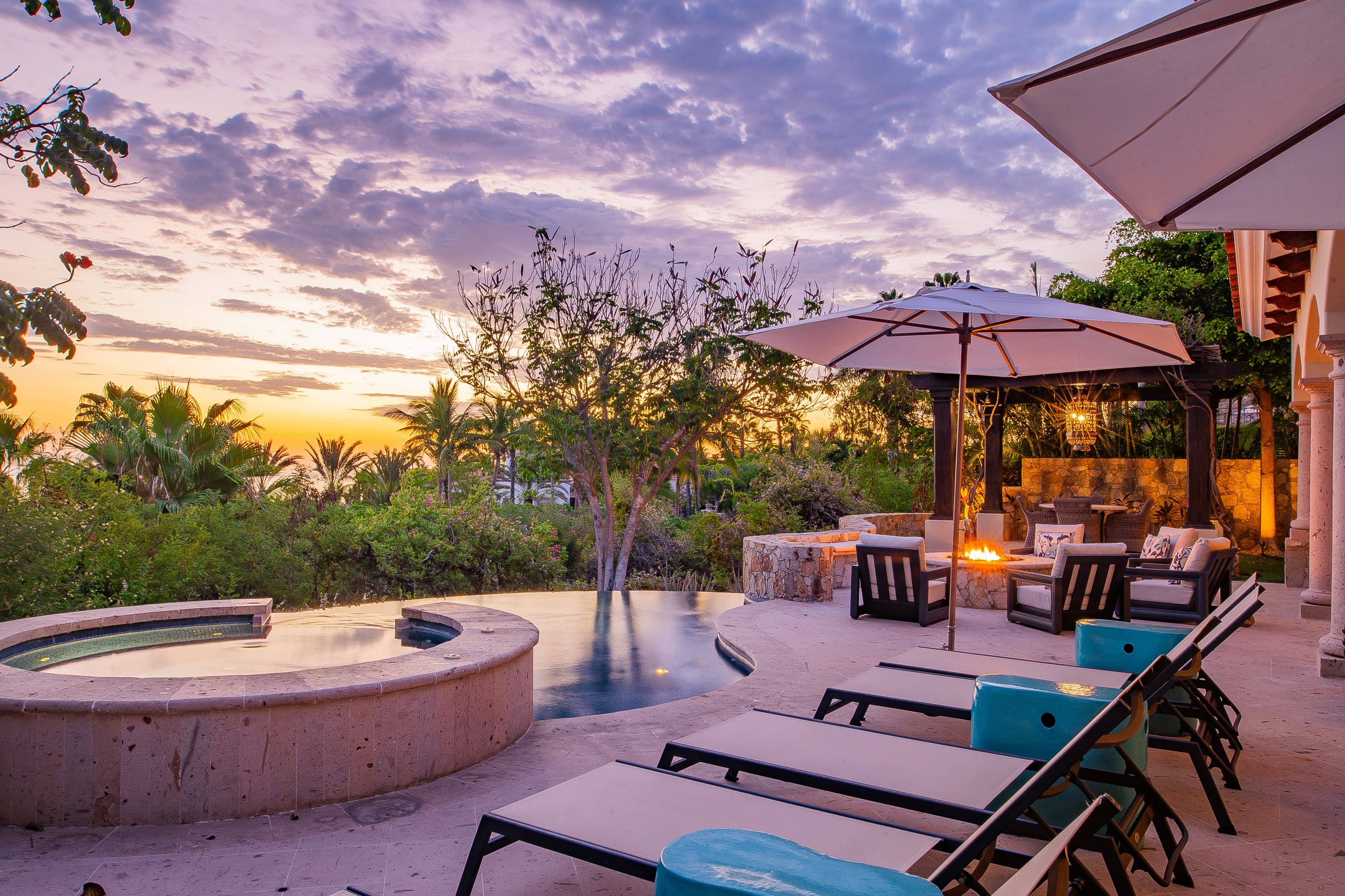 The property offers a serene outdoor setting with a heated pool, patio loungers, and lush tropical landscaping, set against stunning desert sunset views.