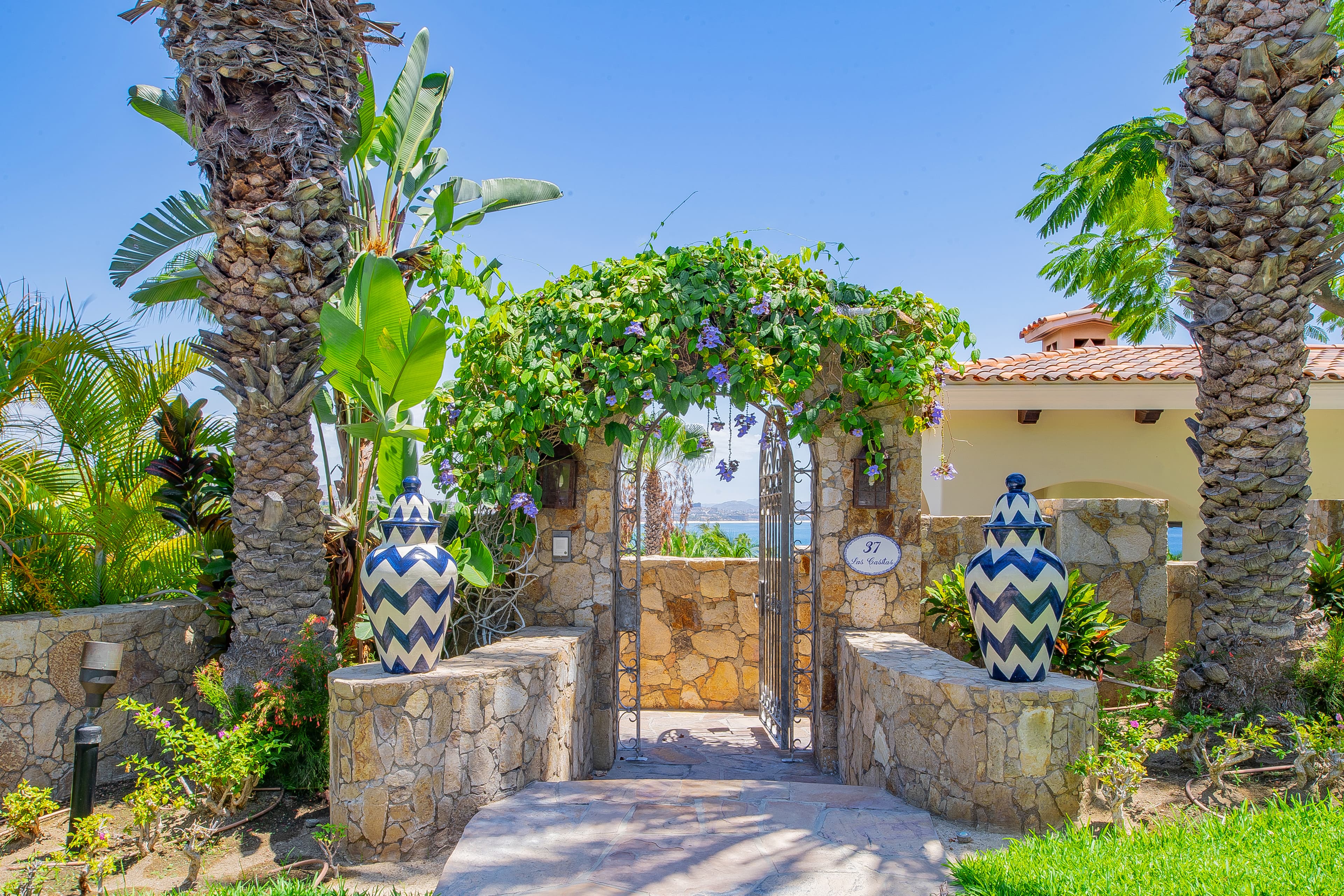 The property entrance features a vine-draped stone archway flanked by tropical palms and decorative blue-and-white vases, welcoming guests to a Mediterranean-inspired coastal retreat.