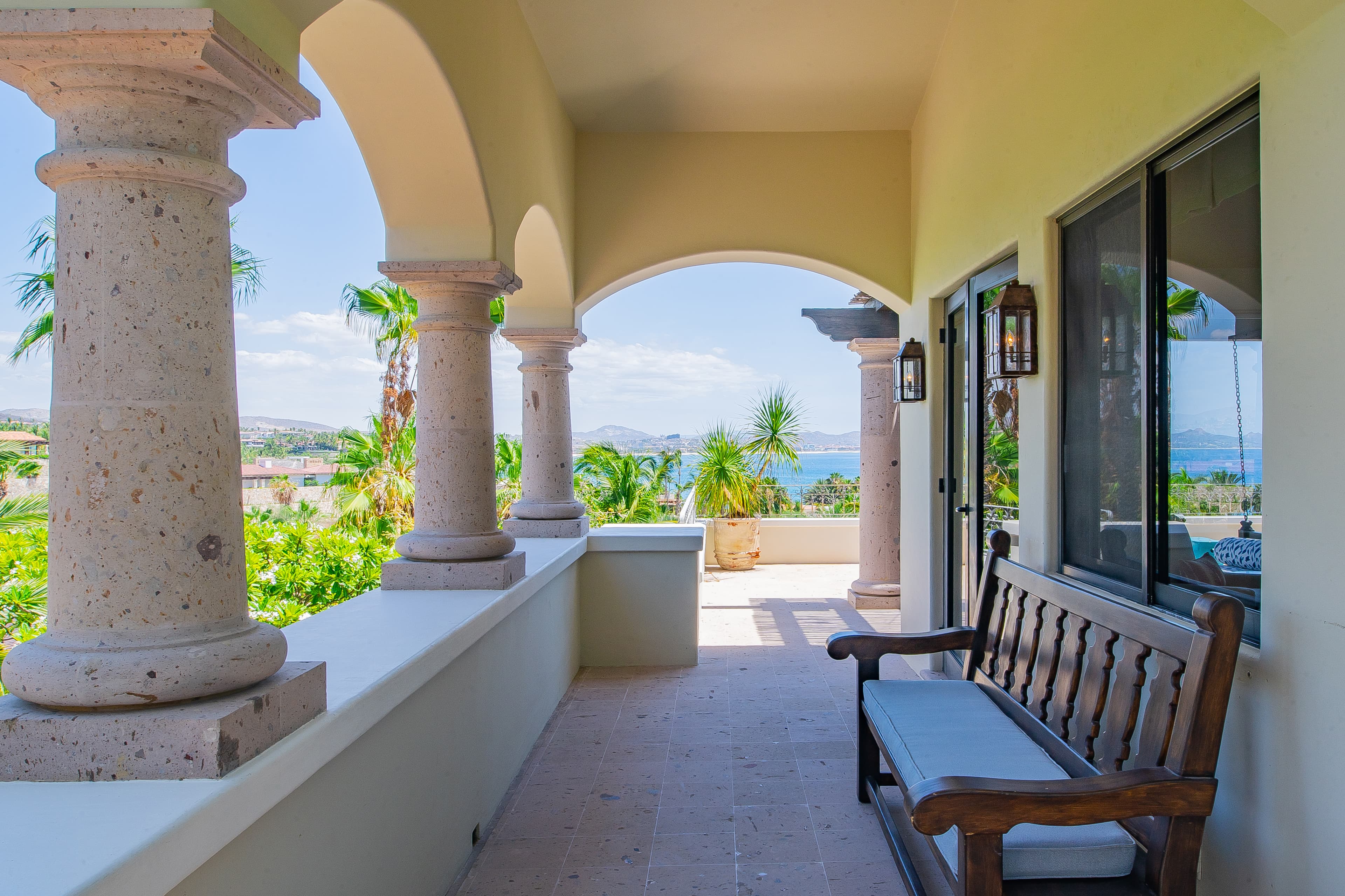 A covered terrace framed by elegant stone columns opens to stunning coastal views. Lush palms and distant blue waters frame this peaceful outdoor sitting area, perfect for relaxing mornings or sunset evenings.
